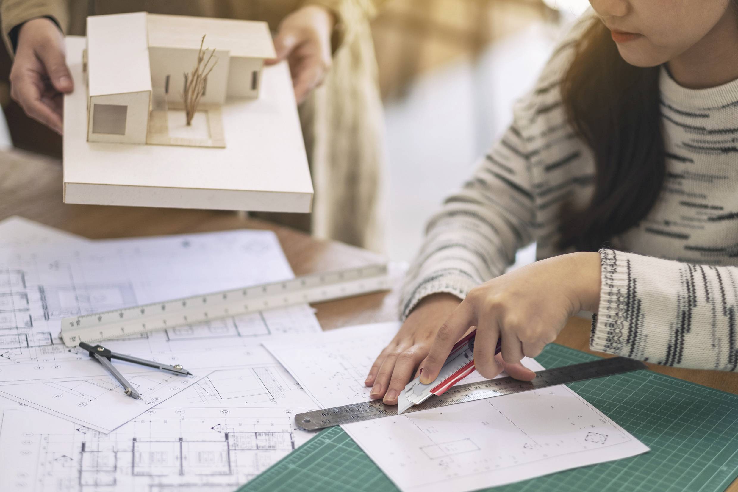 En person ritar på byggritningar medan en annan håller ett arkitekturmodell i händerna.