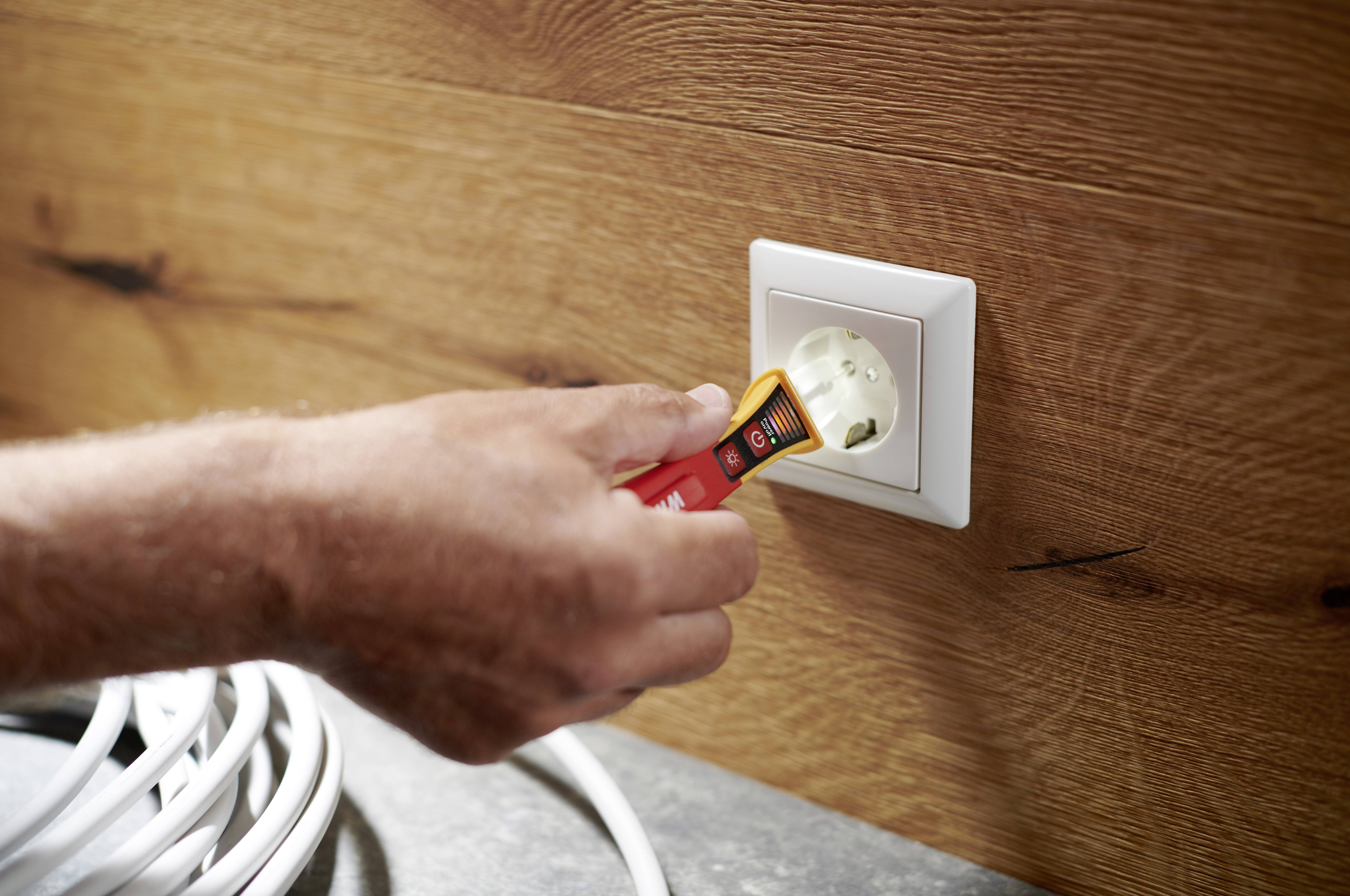 En hand håller en spänningstestare och testar ett vitt vägguttag på en träpanel. En vit kabel ligger bredvid.