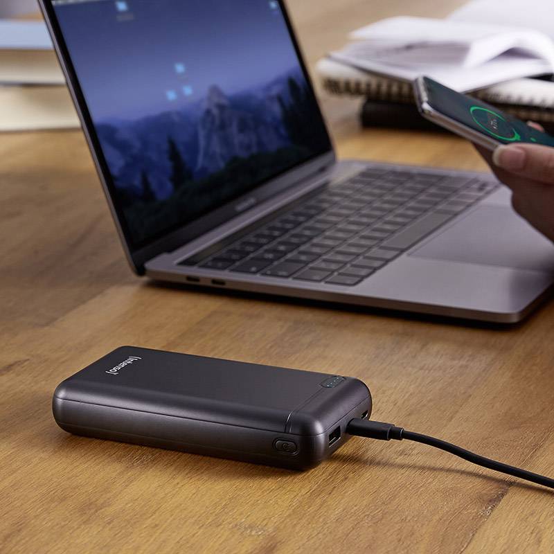 Laptop på ett träbord; bredvid en powerbank som laddar en smartphone. Böcker i bakgrunden.