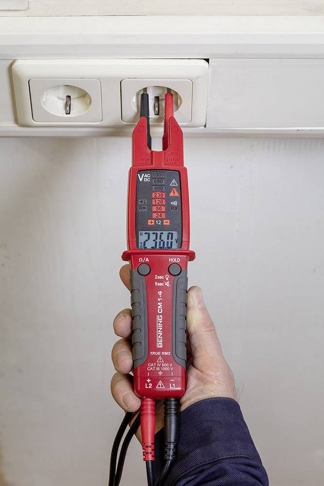 En hand håller ett digitalt multimeter som är anslutet till ett vägguttag. Displayen visar '230,0 V', vilket representerar den uppmätta spänningen.