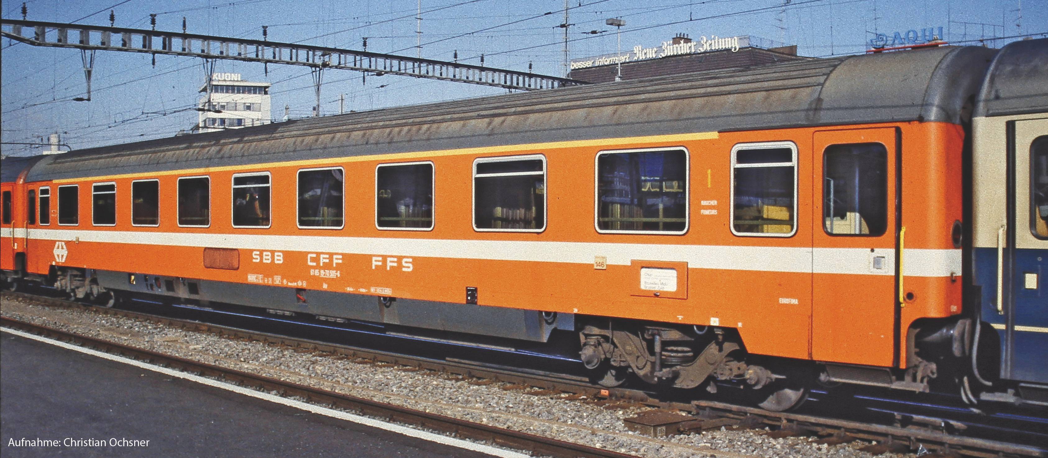 Ett orange tåg från SBB står på ett spår vid järnvägsstationen. I bakgrunden syns kontaktledningar, byggnader och en företagslogotyp.