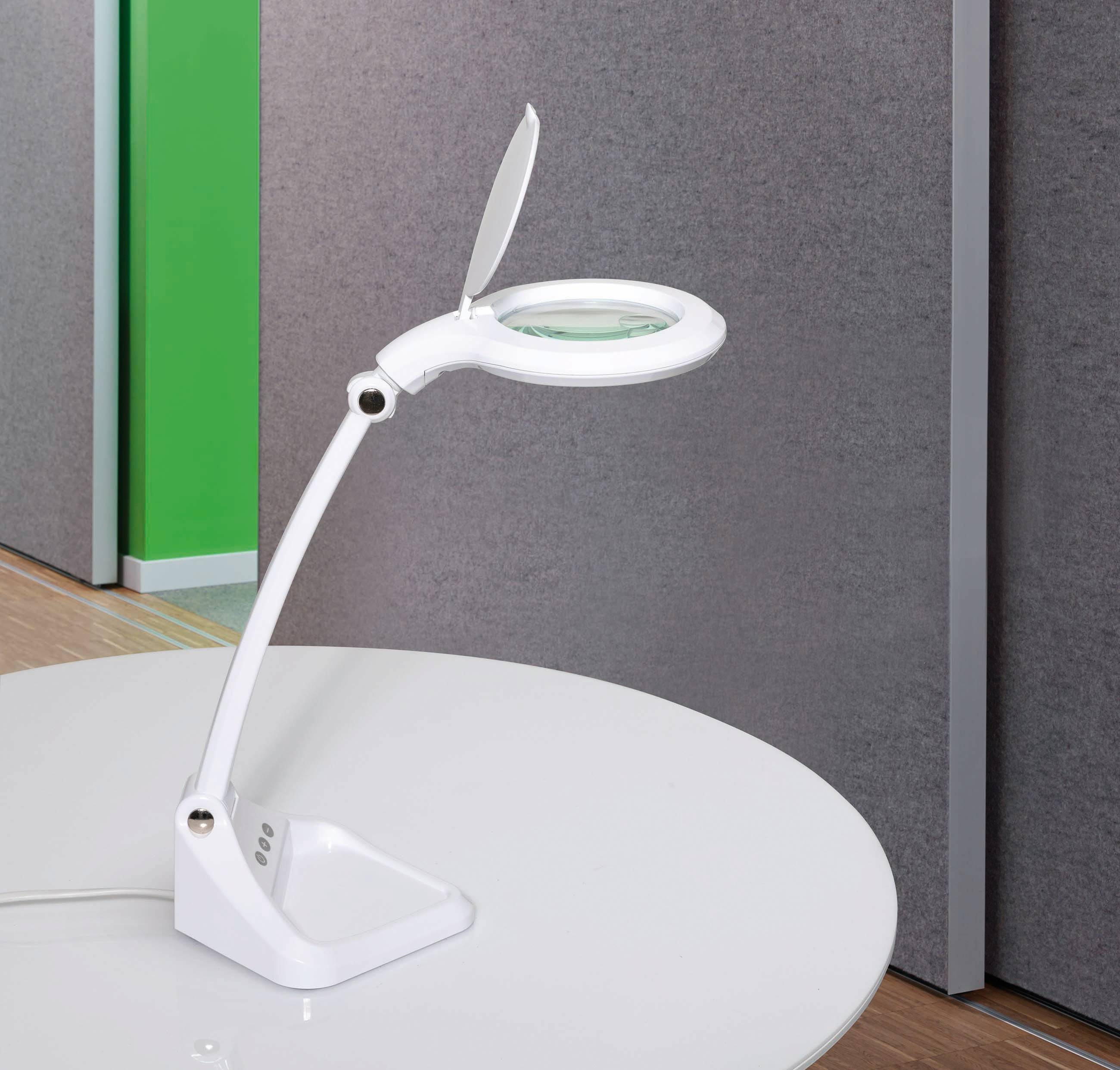 En vit bordslampa med justerbar arm och lupp står på ett vitt bord framför en grå-grön vägg.