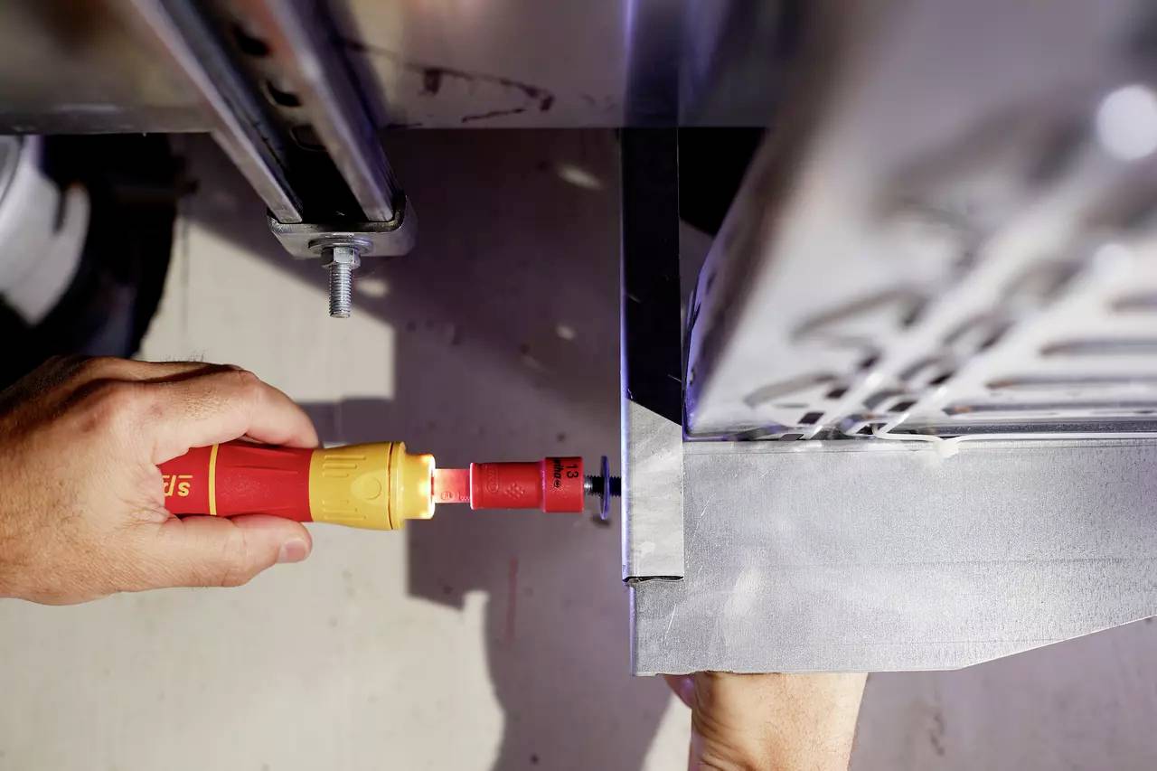 En hand med skruvmejsel fäster en metallplatta vid en ram. Verktyg i aktion nära en maskinstruktur.