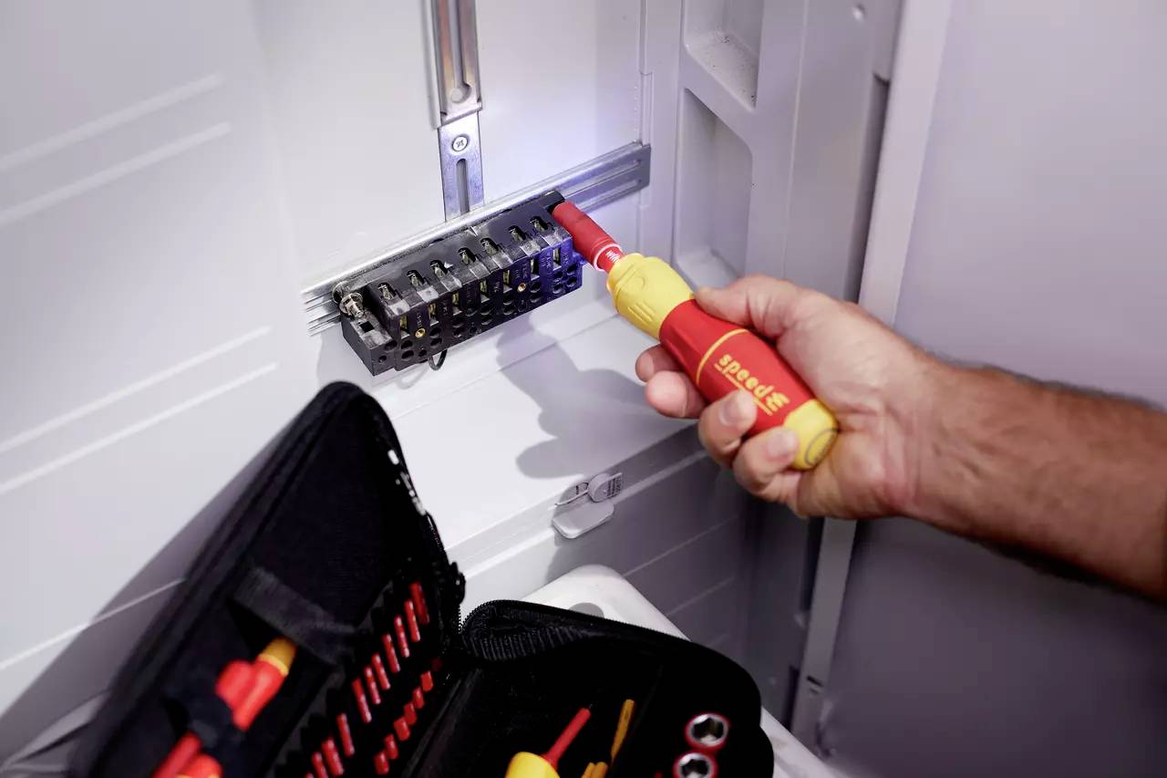 En hand använder en isolerad skruvmejsel för att arbeta vid en elektrisk kopplingstavla. En verktygslåda ligger intill.
