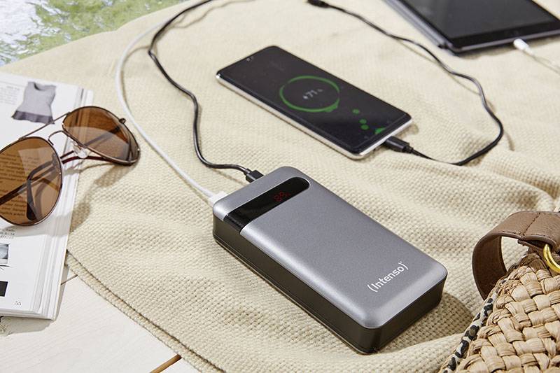 Bärbar powerbank laddar smartphonen. En surfplatta, solglasögon och en öppen tidskrift ligger bredvid på en filt.