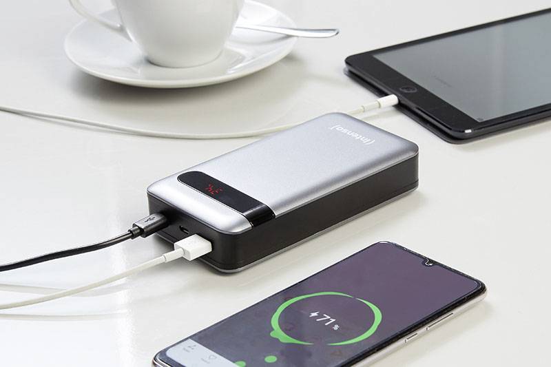 En powerbank laddar två enheter på ett bord: en smartphone med 71% batterinivå och en surfplatta. I bakgrunden en kopp.