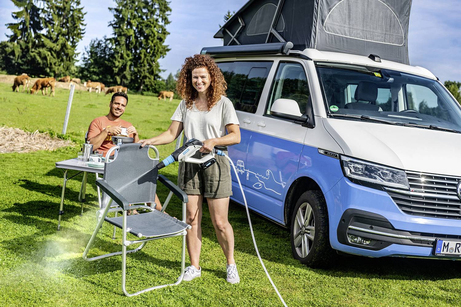 En man sitter på en campingstol och dricker kaffe bredvid en blå husbil; i bakgrunden betar kor på en äng.