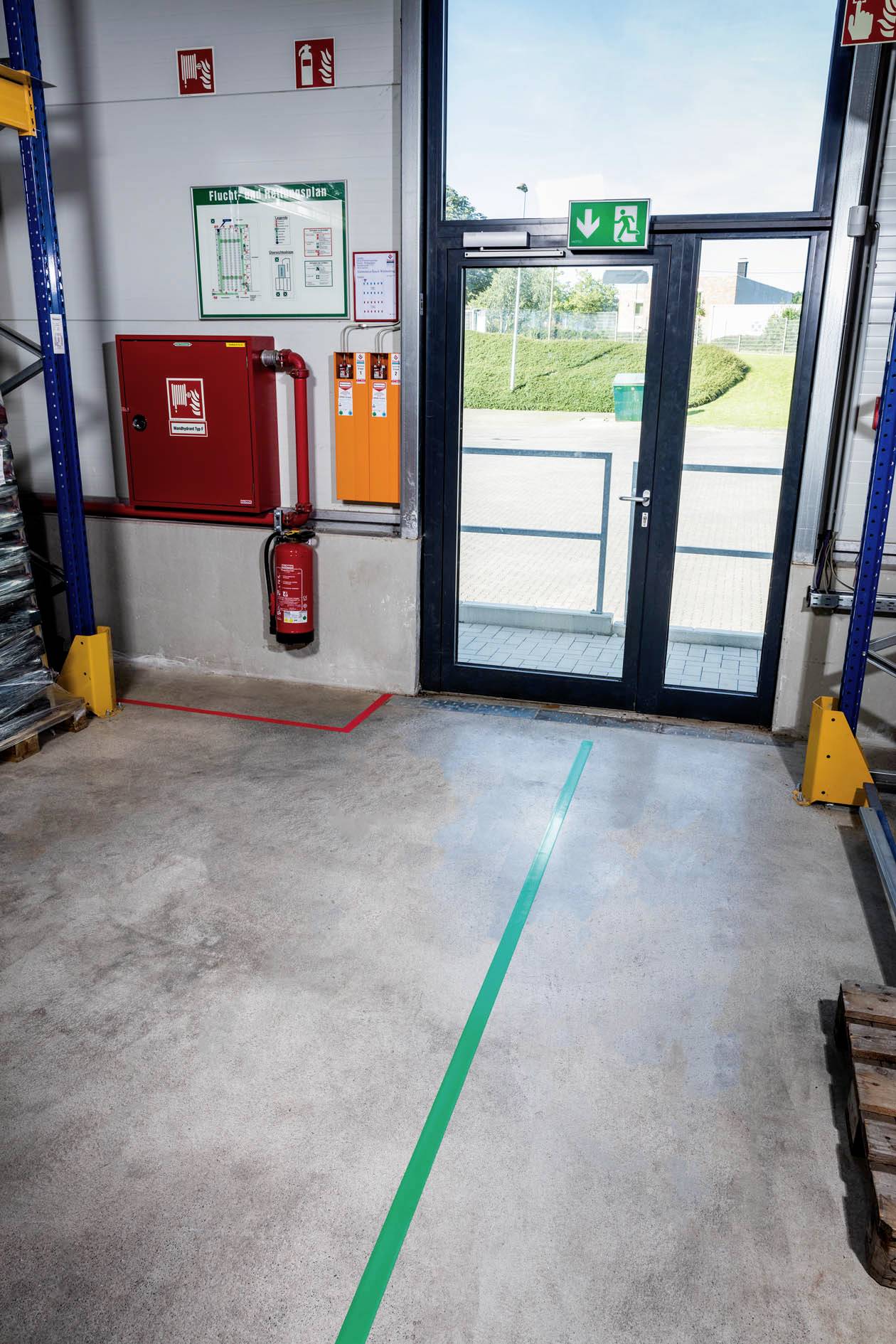 En nödutgångsdörr i ett lager med utrymningsmarkeringar på golvet, brandsläckare, nödplan och säkerhetsskylt på väggen.