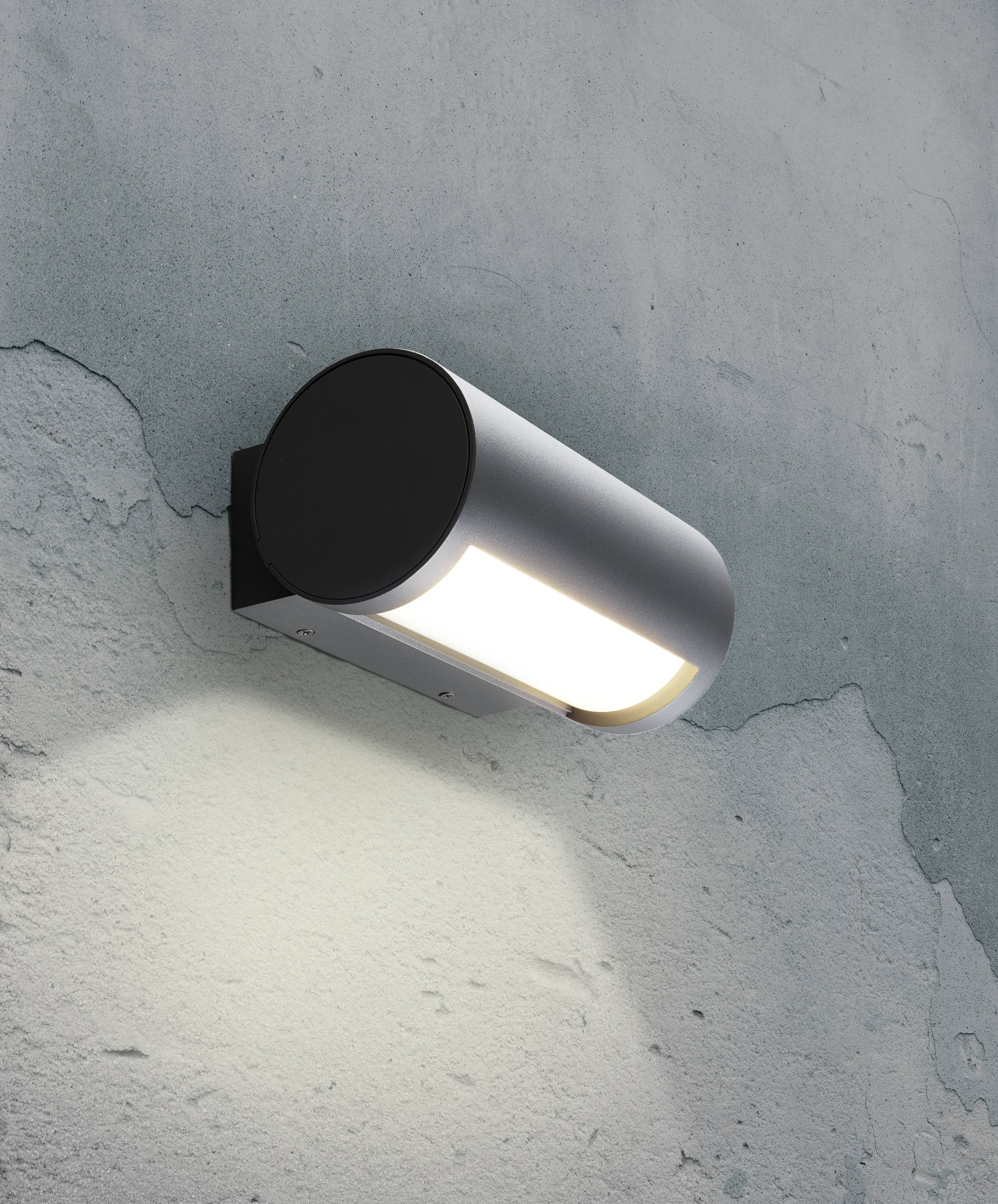 En modern, cylindrisk vägglampa med svart fäste kastar ett varmt ljus på en gröngrå, grov väggyta.