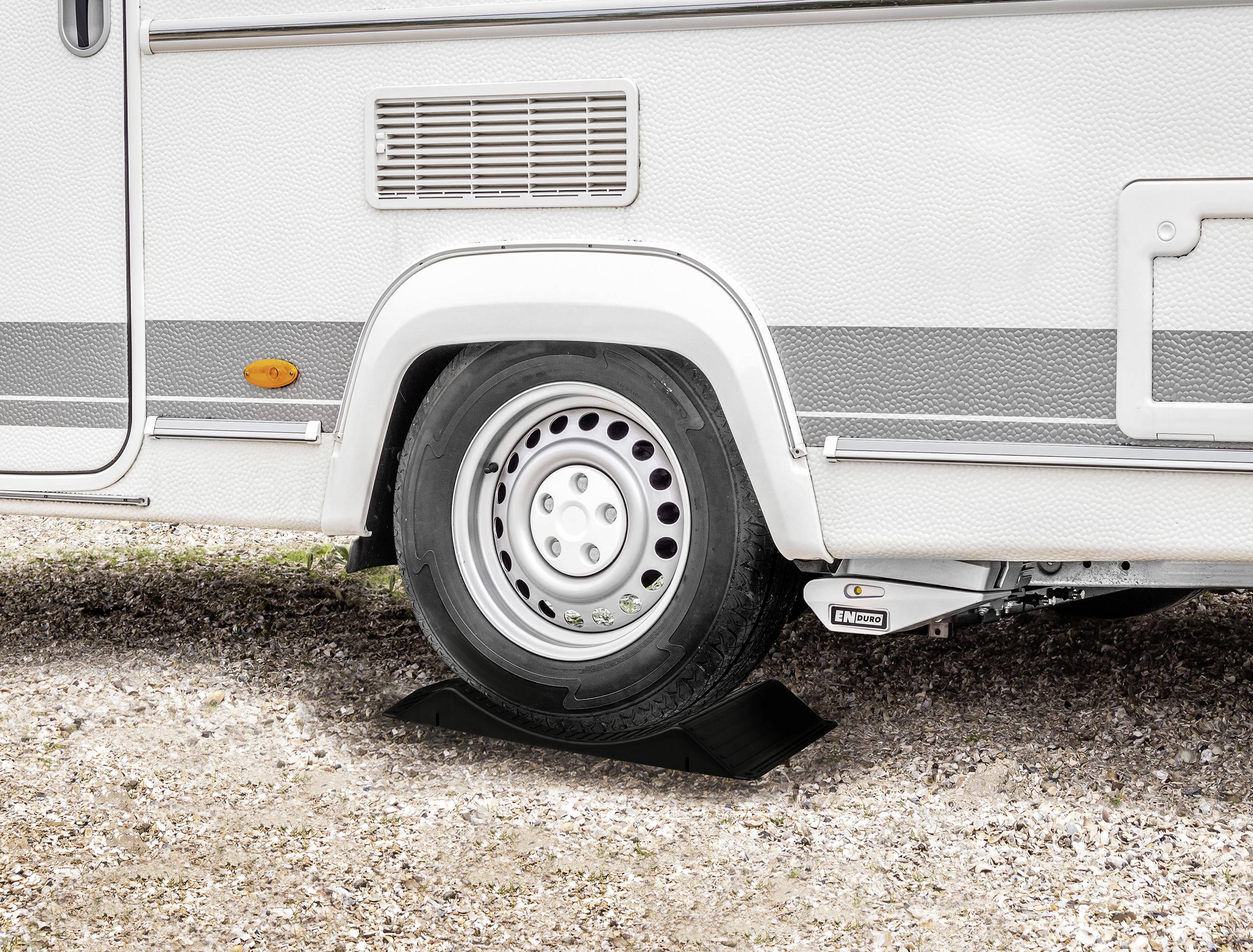 Husbil, parkerad på grusväg, med ett hjul på en svart kil för stabilitet och nivellering.