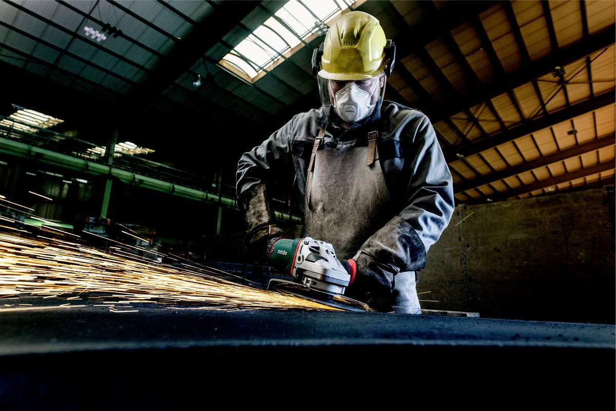 En arbetare med skyddshjälm och skyddsglasögon sliper metall i en industrihall. Gnistor sprutar från verktyget.