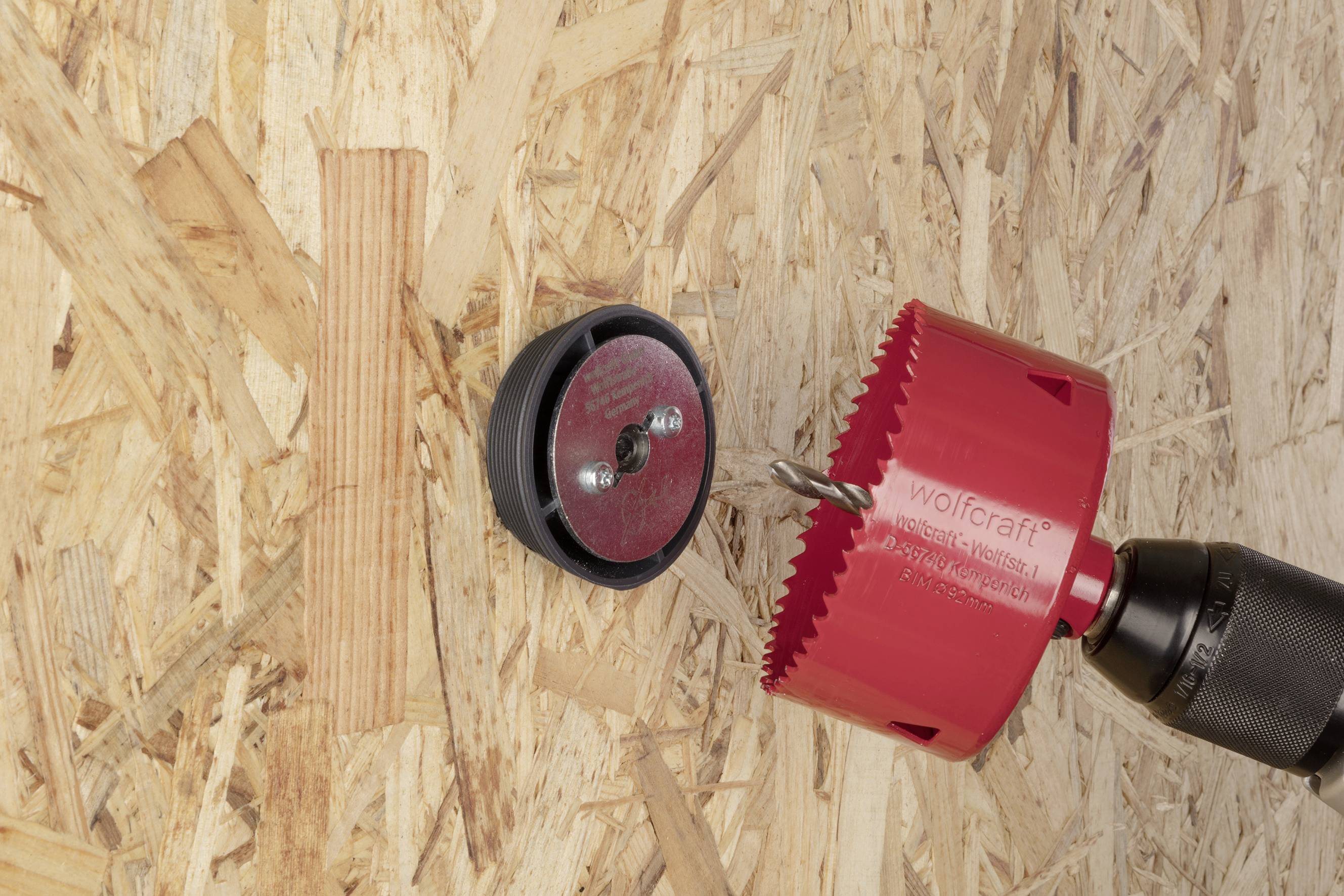 En röd hålsågsadapter på en bormaskin borrar ett hål i en spånskiva. En svart anslagsring är fäst vid brädan.