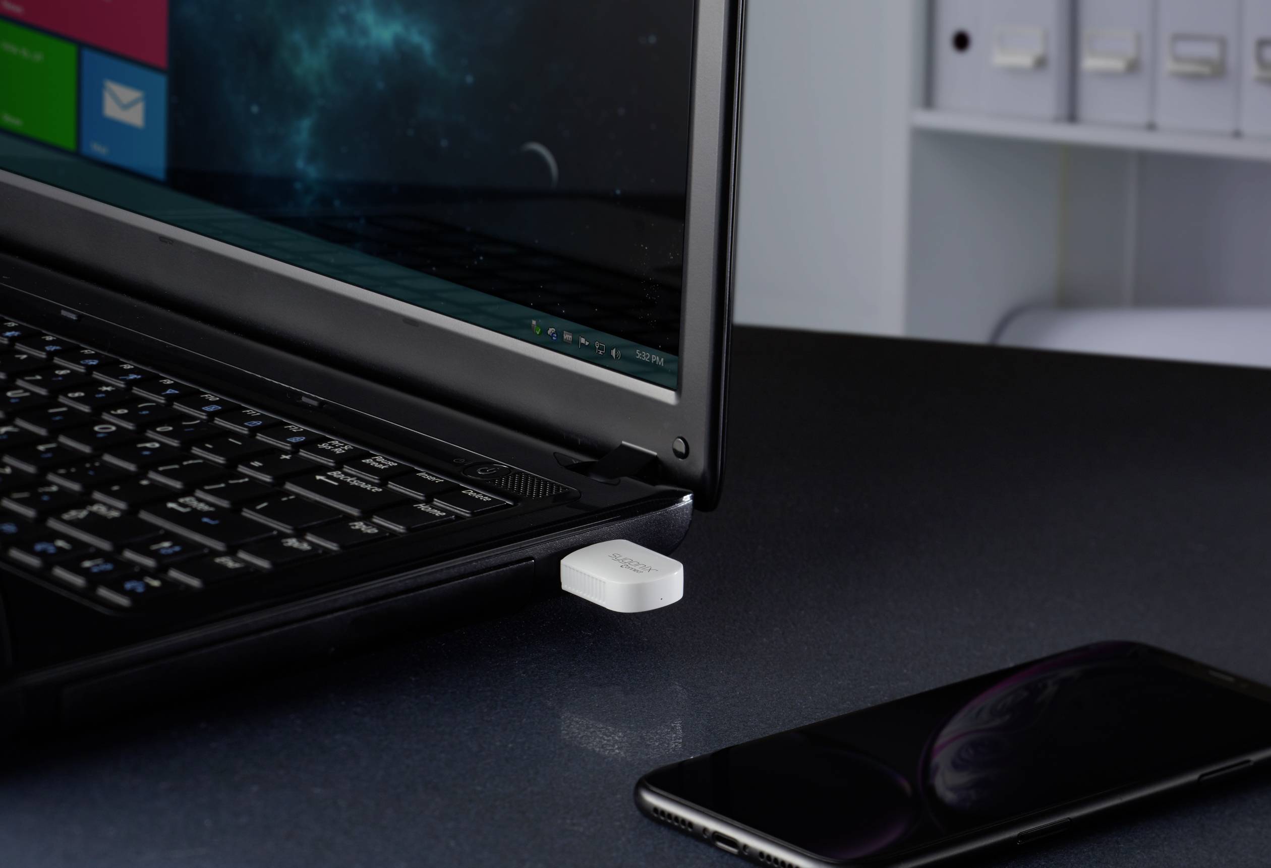 En laptop står på ett bord, en USB-dongle är isatt. Bredvid ligger en smartphone. I bakgrunden syns pärmar.