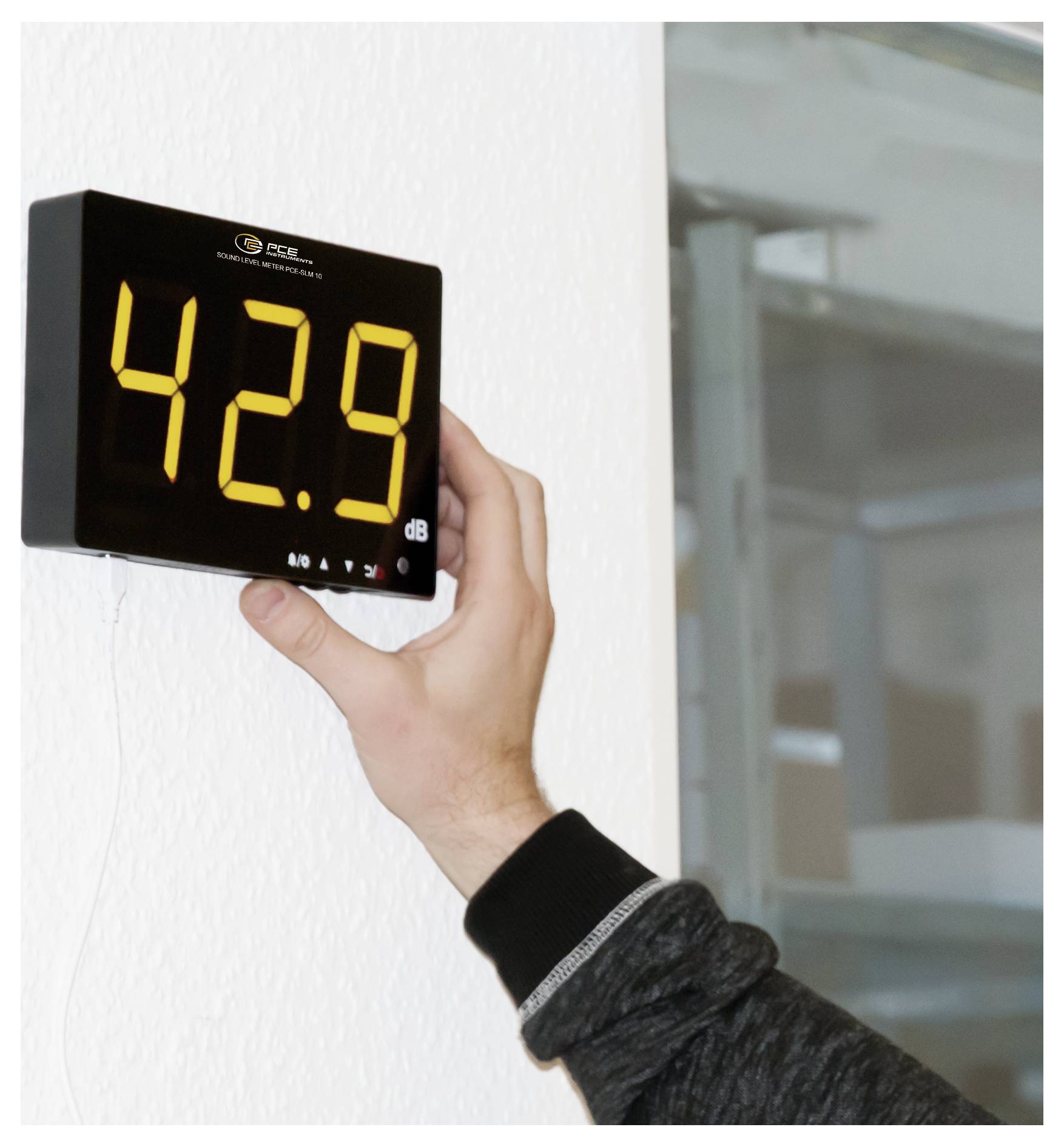Hand som håller en digital ljudnivåmätare monterad på en vägg, som visar 42,9 dB.