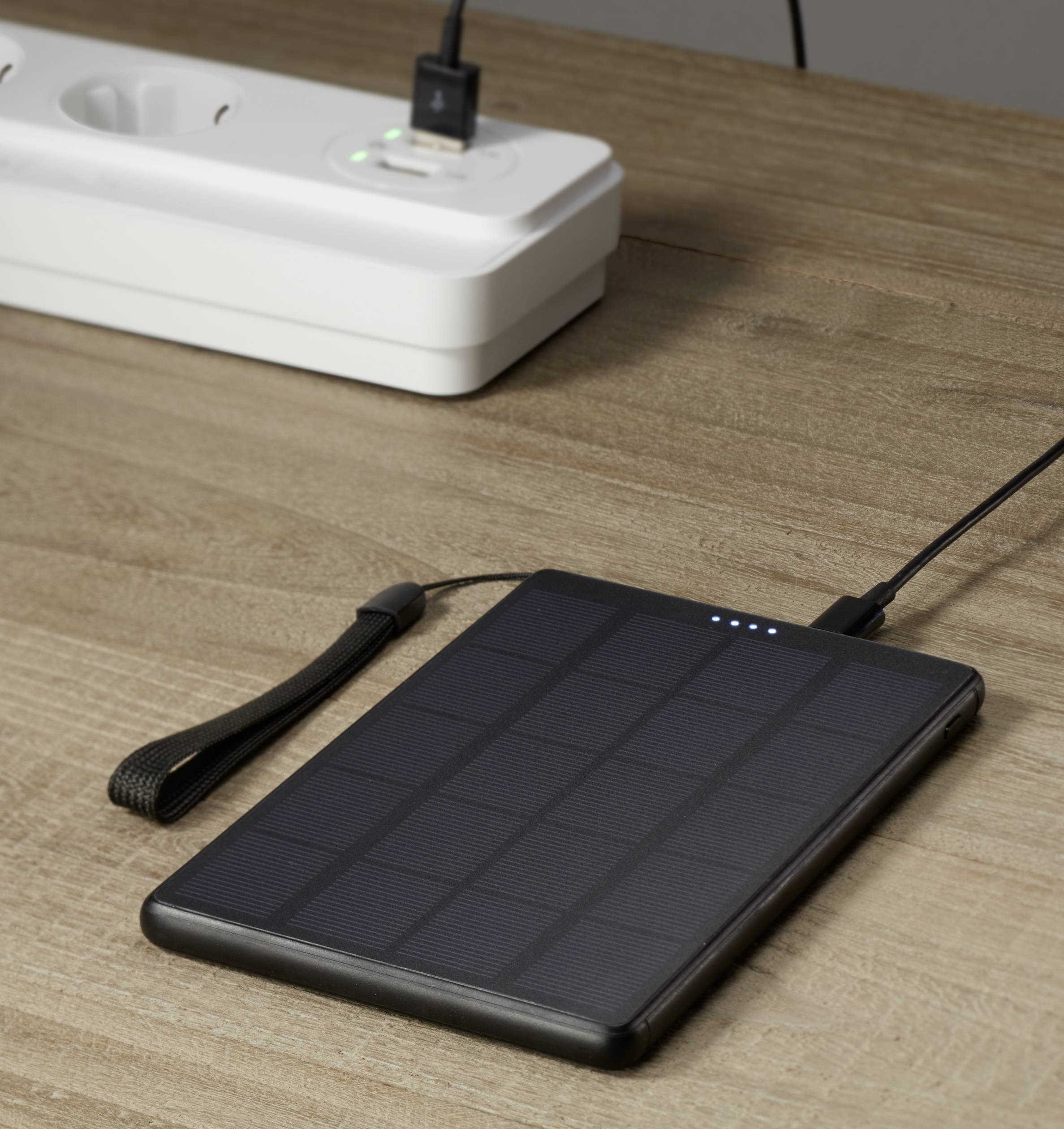 Solcellsdriven powerbank med laddningsindikator, liggande på ett träbord, ansluten via en kabel till ett grenuttag.