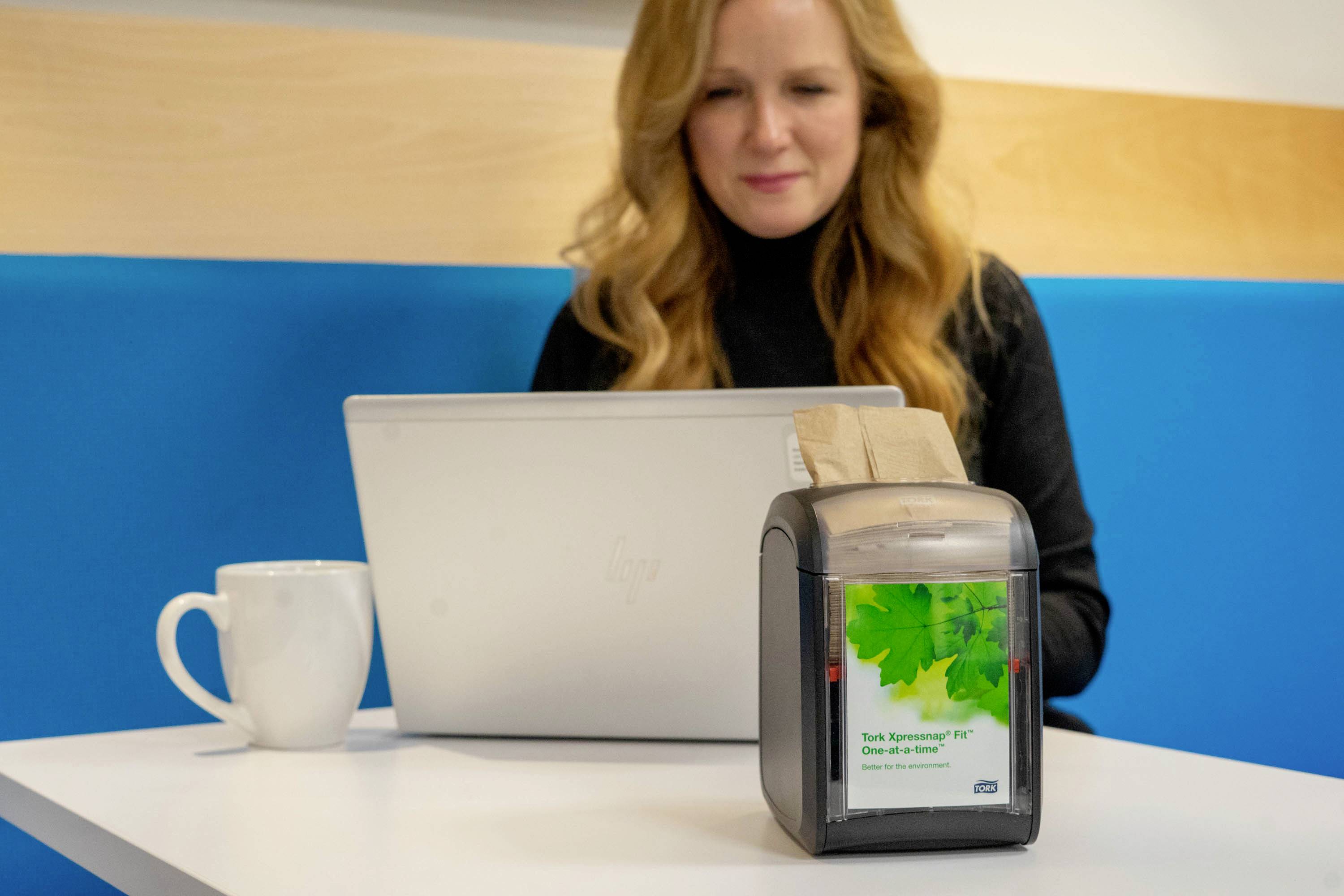 En kvinna med långa, blonda hår arbetar på en bärbar dator på ett café. På bordet står en pappersduk och en vit kopp.