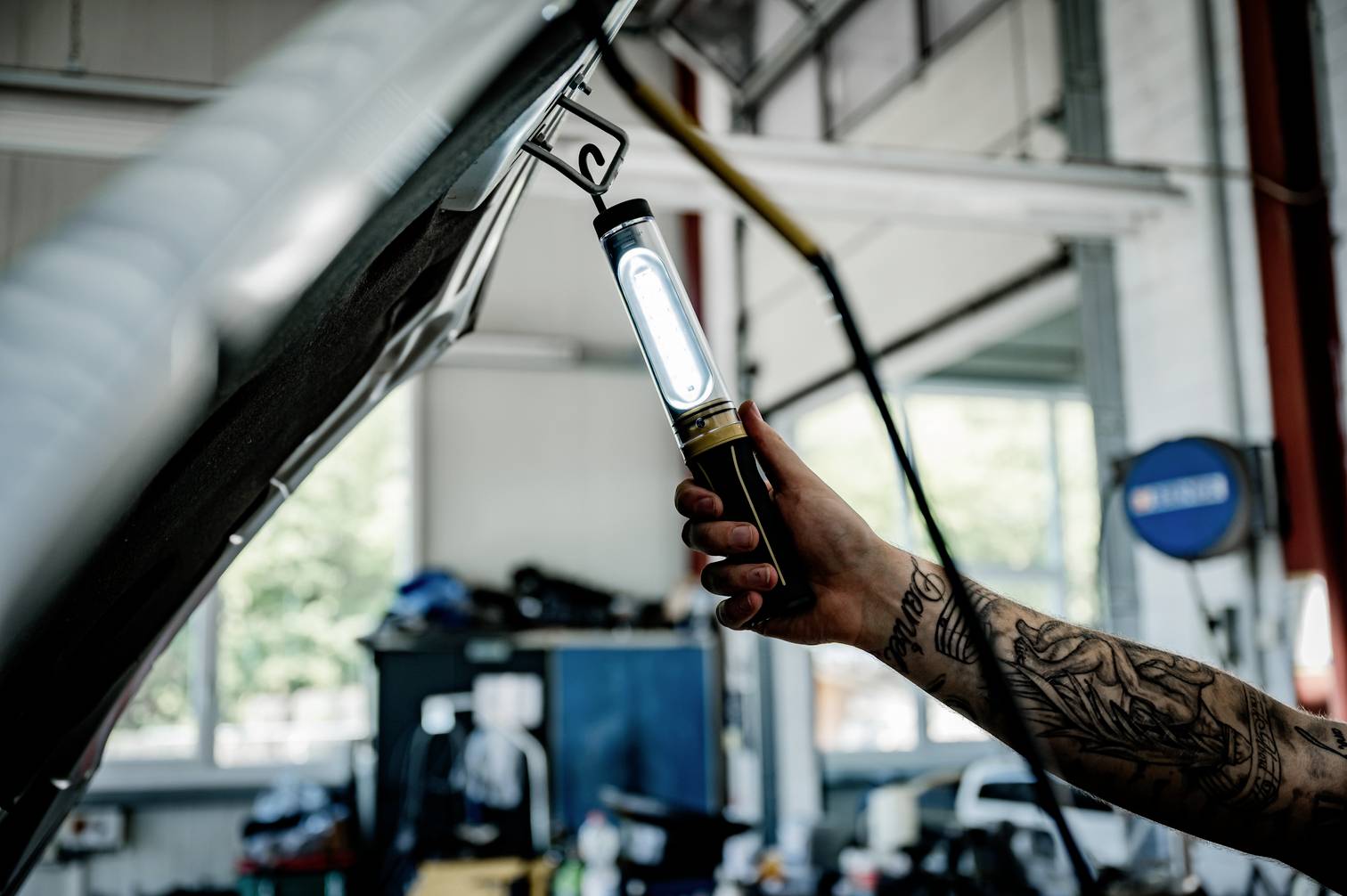 En tatuerad arm håller en arbetslampa under en öppen motorhuv i en bilverkstad. I bakgrunden syns verktyg.