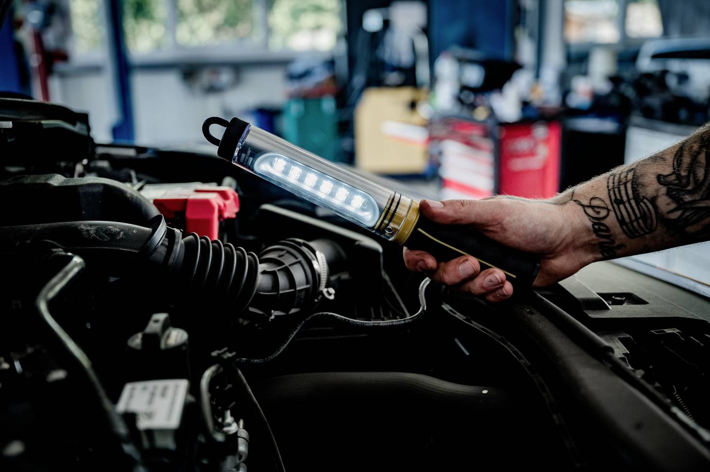 En person håller en verkstadslampa över ett öppet motorrum i en bilverkstad.