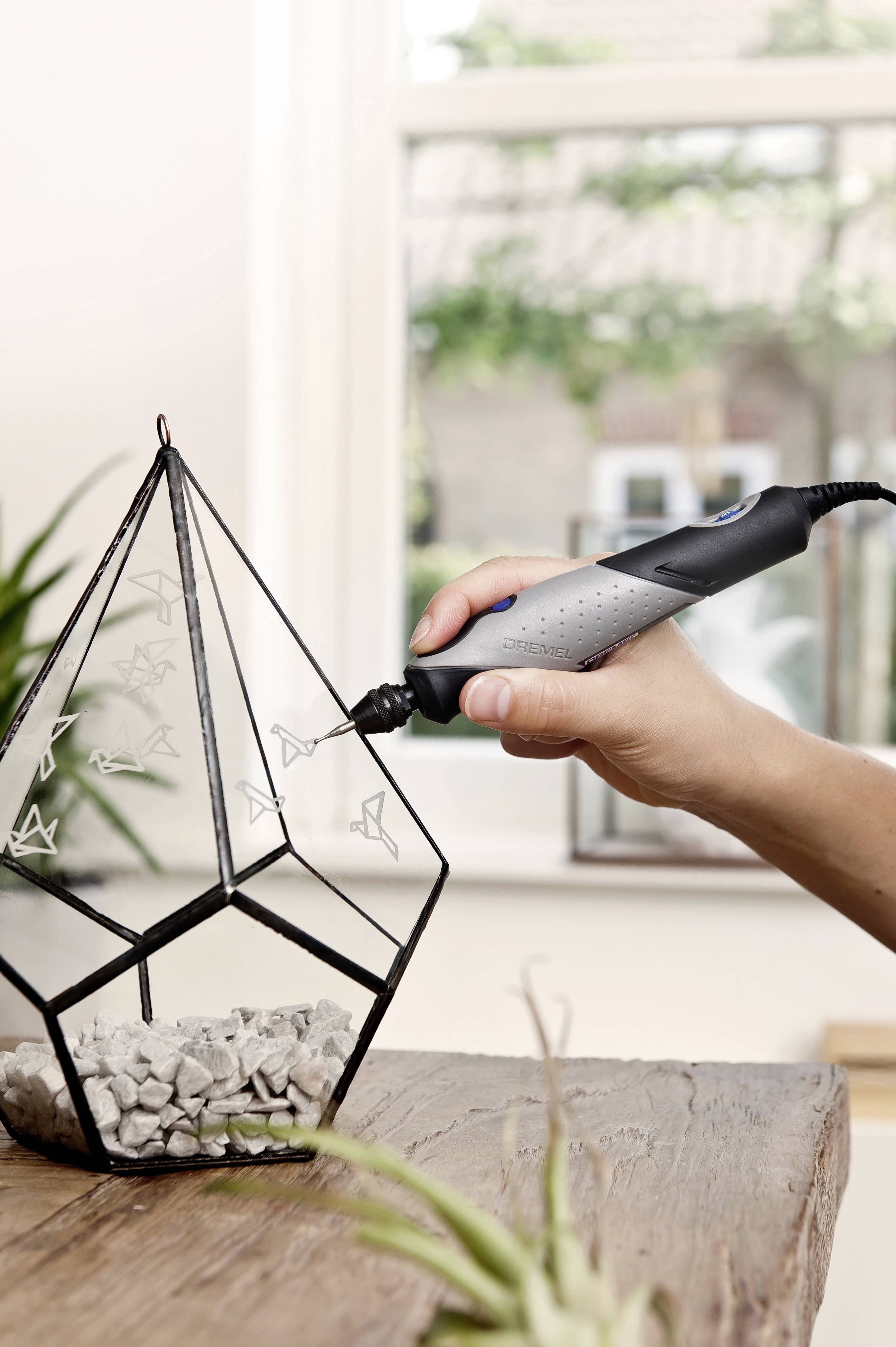 En hand graverar geometriska mönster på en glasvas med ett elektroniskt verktyg. I bakgrunden syns fönster.