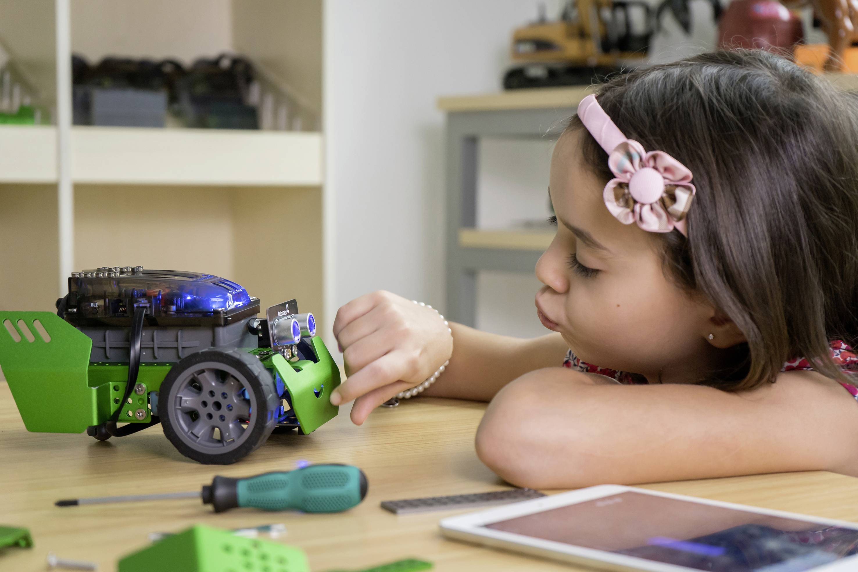 Ett barn leker med en liten, grön robot på ett bord. Det ser intresserat ut medan det justerar en del av roboten. I bakgrunden syns hyllor med fler leksaker.