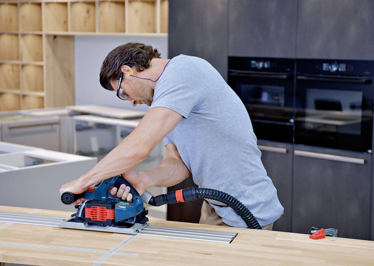 En man sågar trä med en elsåg i en verkstad. Han bär skyddsglasögon och hörselskydd.