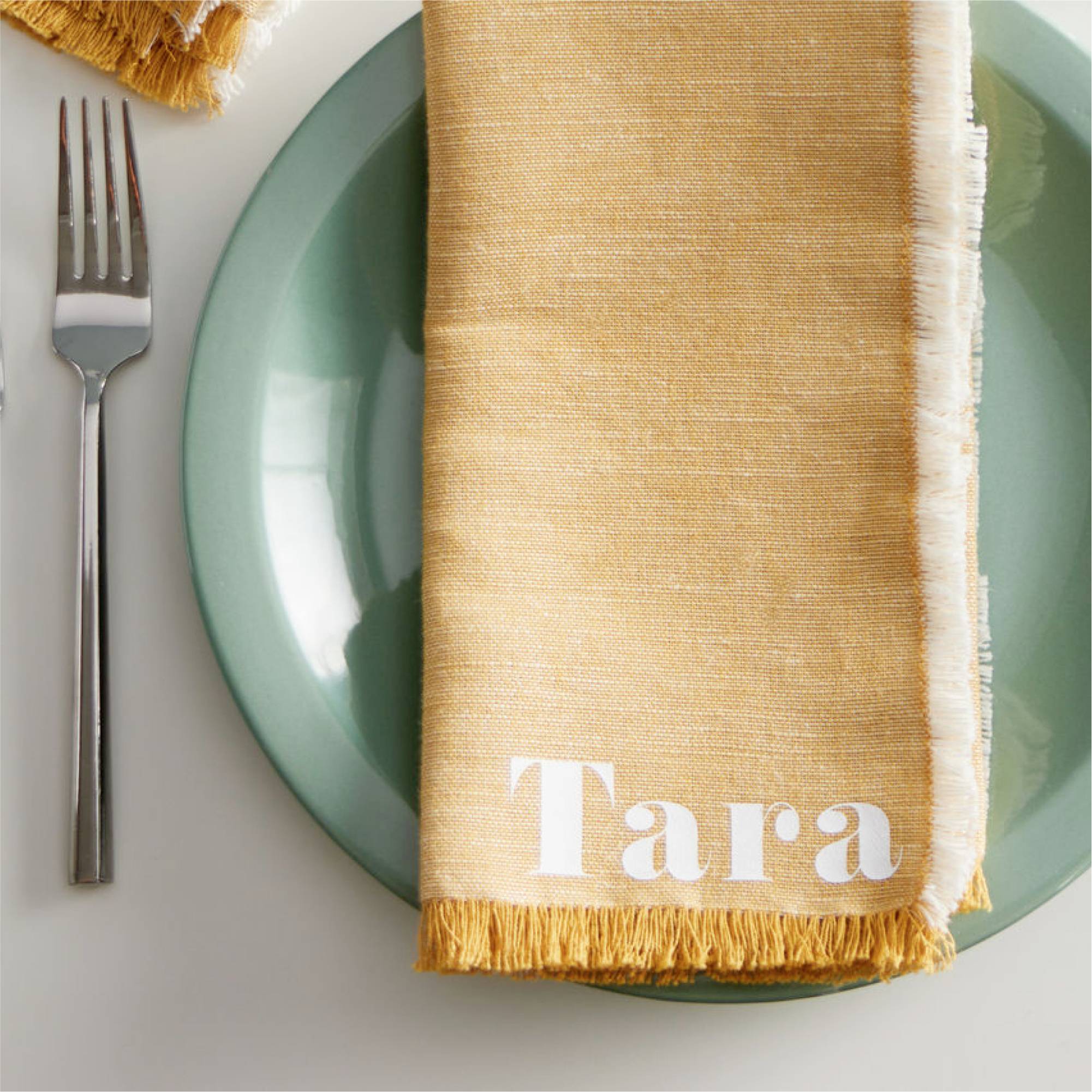 Gul tygservett med namnet 'Tara' på en grön tallrik, bredvid en gaffel.