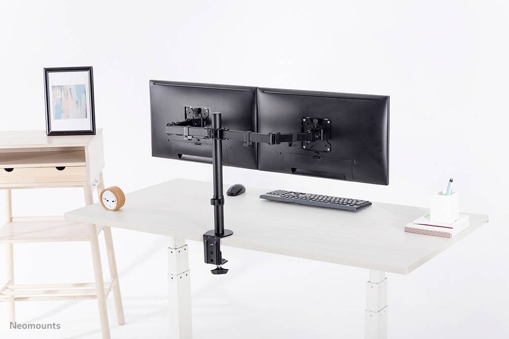 Ett vitt skrivbord med två skärmar på ett flexibelt fäste. Bredvid ett litet bord med dekorationer.