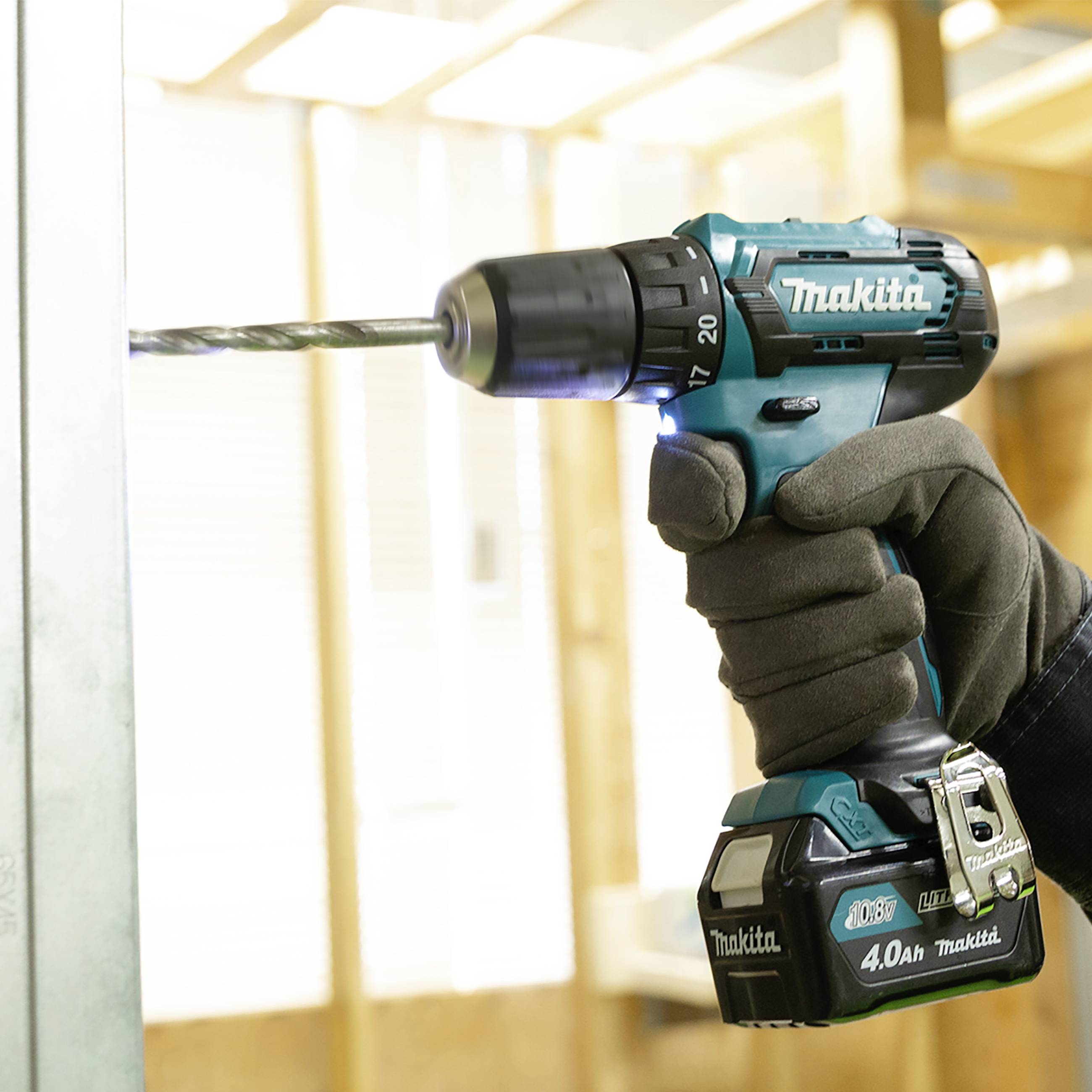 En hand borrar med en blå Makita-borr i en trävägg. I bakgrunden syns oskarpa träbalkar.