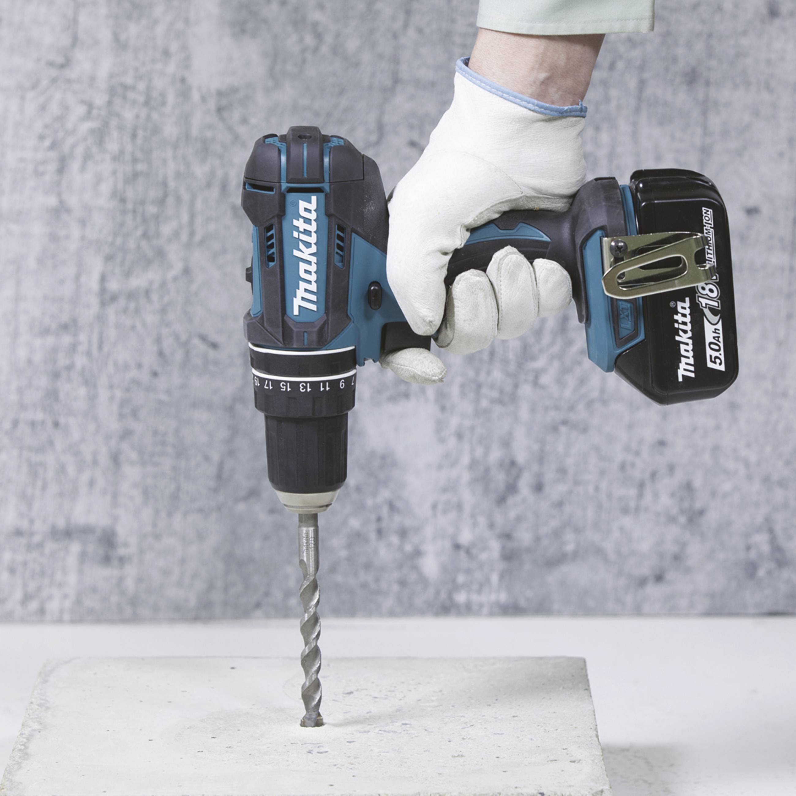 Borrmaskin, hållen av en handskklädd hand, borrar i betongyta. Märket 'Makita' och modellnummer synligt.