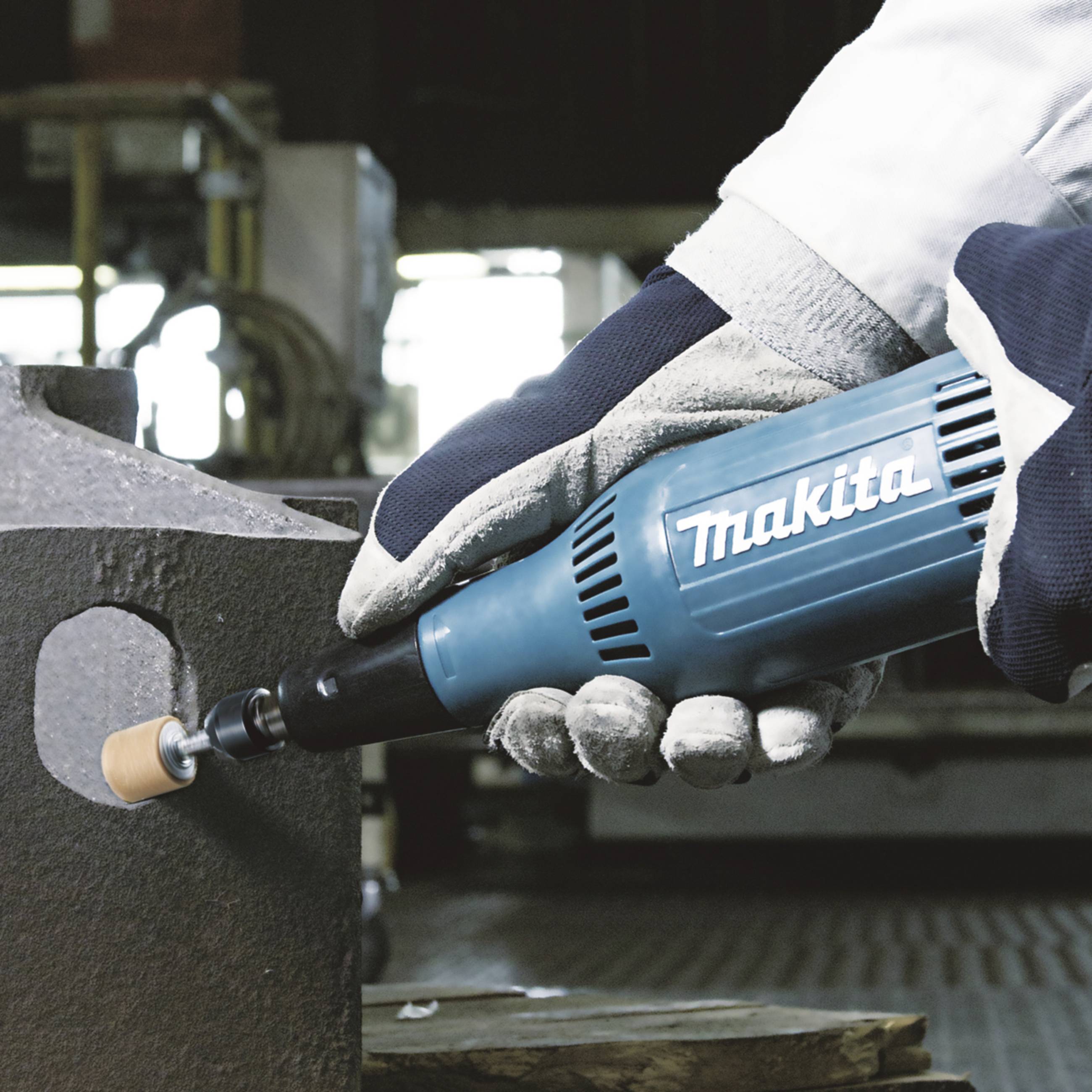 Arbetare med skyddshandskar sliper metall med ett blått Makita-verktyg; industriell miljö i bakgrunden.