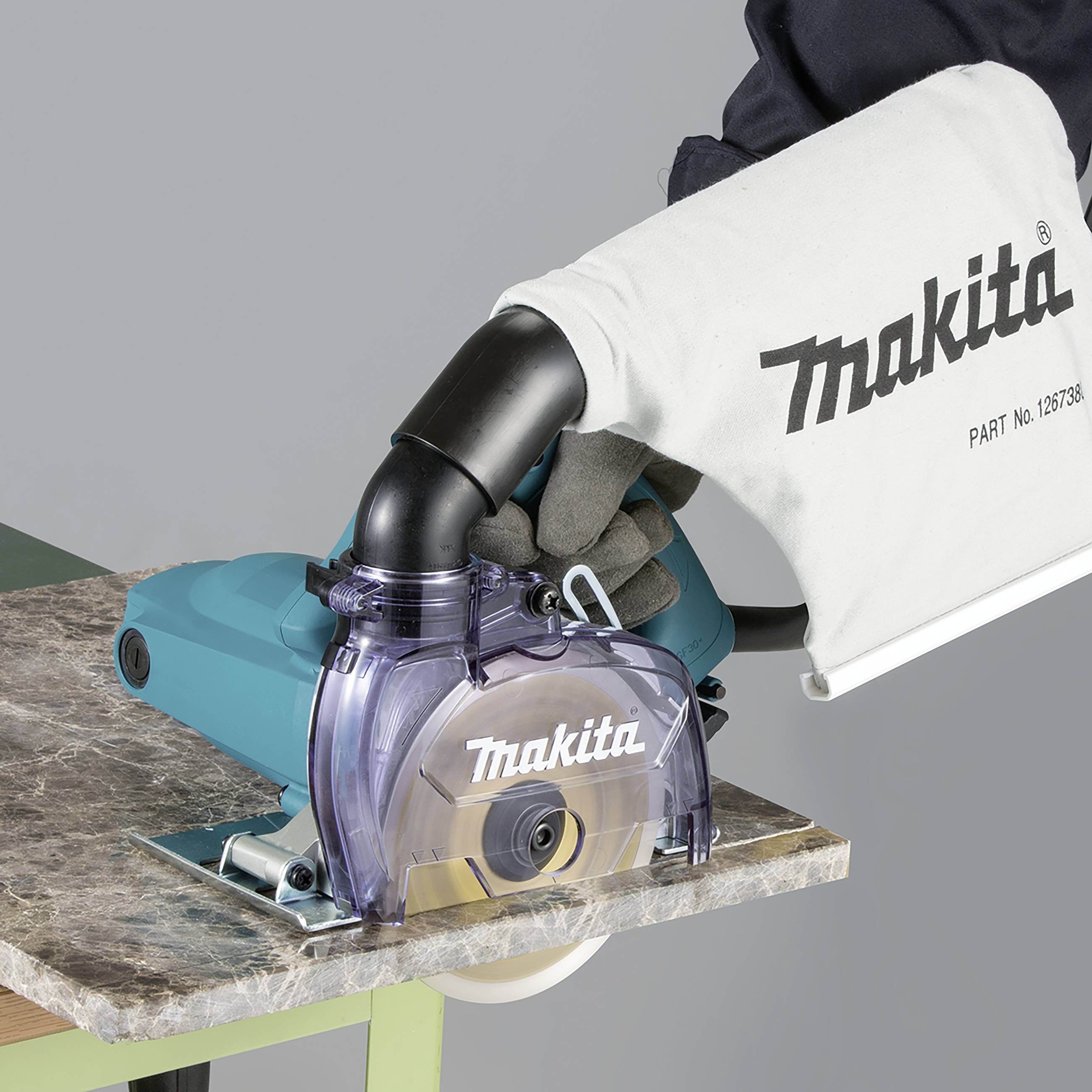 En hand håller en Makita-cirkelsåg med dammpåse, placerad på ett träbord.