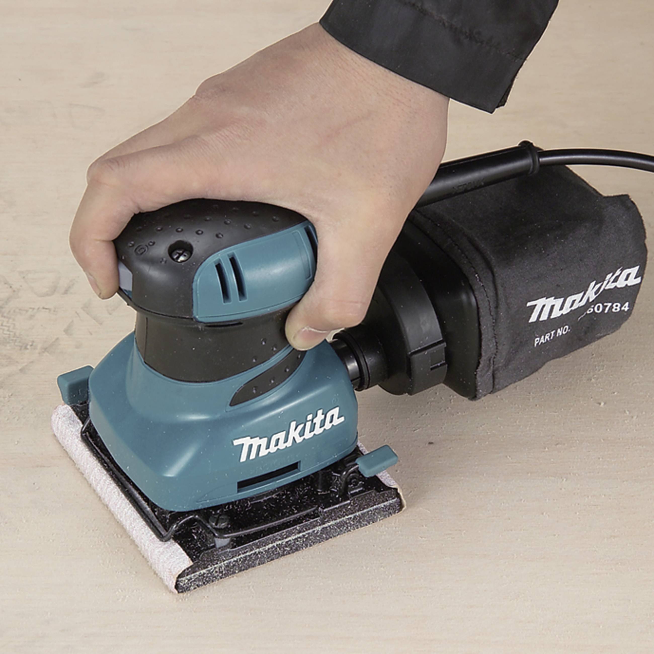 En hand manövrerar en blå Makita-slipmaskin på en träyta. Slipmaskinen är påslagen och jämnar ut ytan.