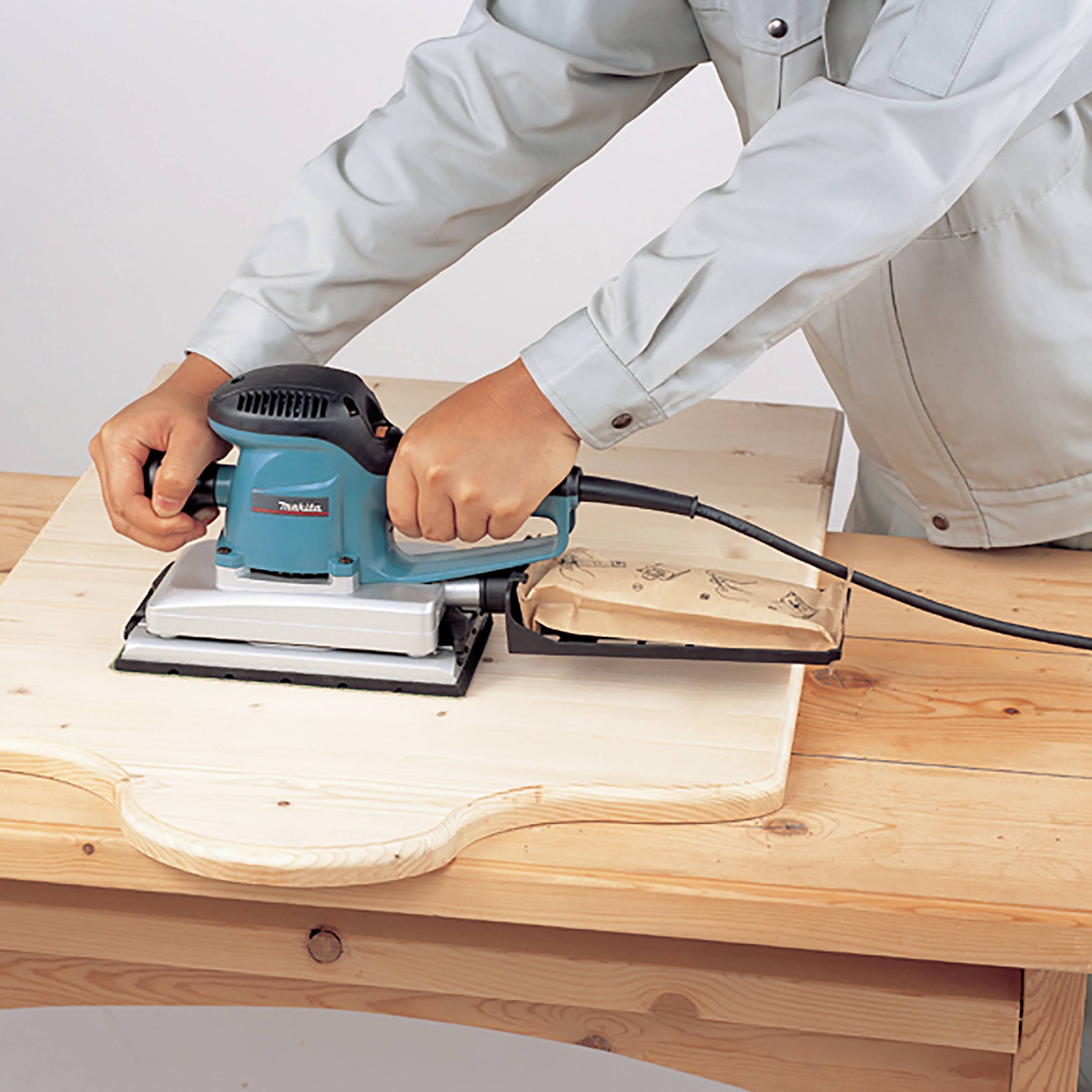 En person sliper ett träbräde med en elektrisk slipmaskin på ett träbord.