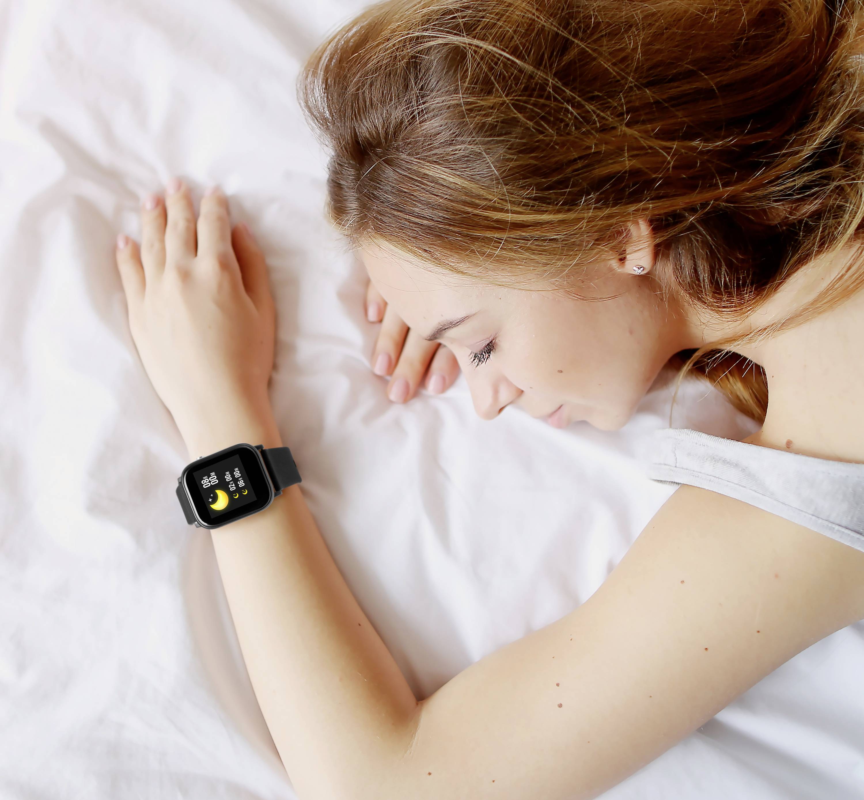 En kvinna ligger sovande i sängen och bär en smartwatch på handleden; klockan visar sömnövervakningsfunktioner.