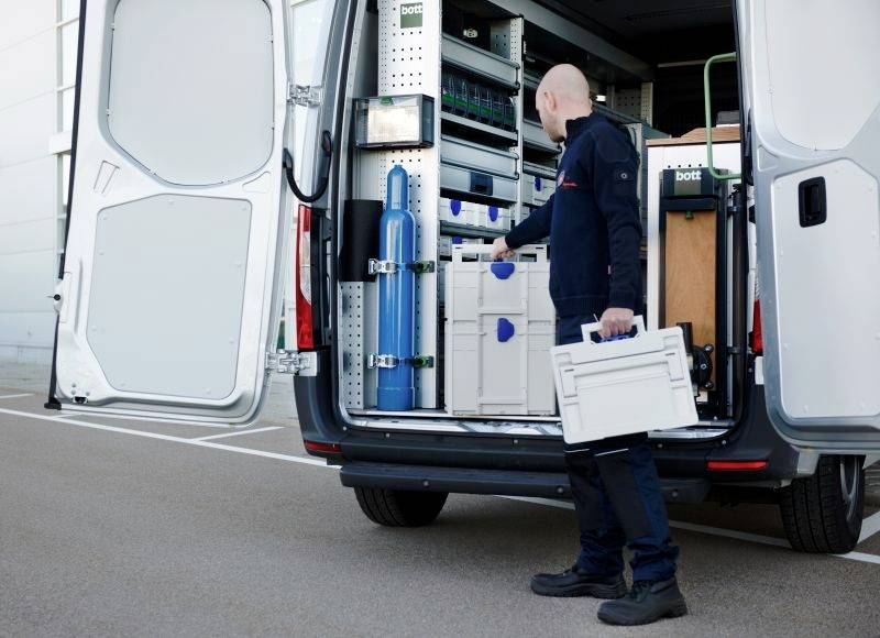 En person organiserar bärbara förvaringsenheter inuti en öppen skåpbil fylld med diverse utrustning, vilket antyder en arbetsrelaterad uppgift eller förberedelse.