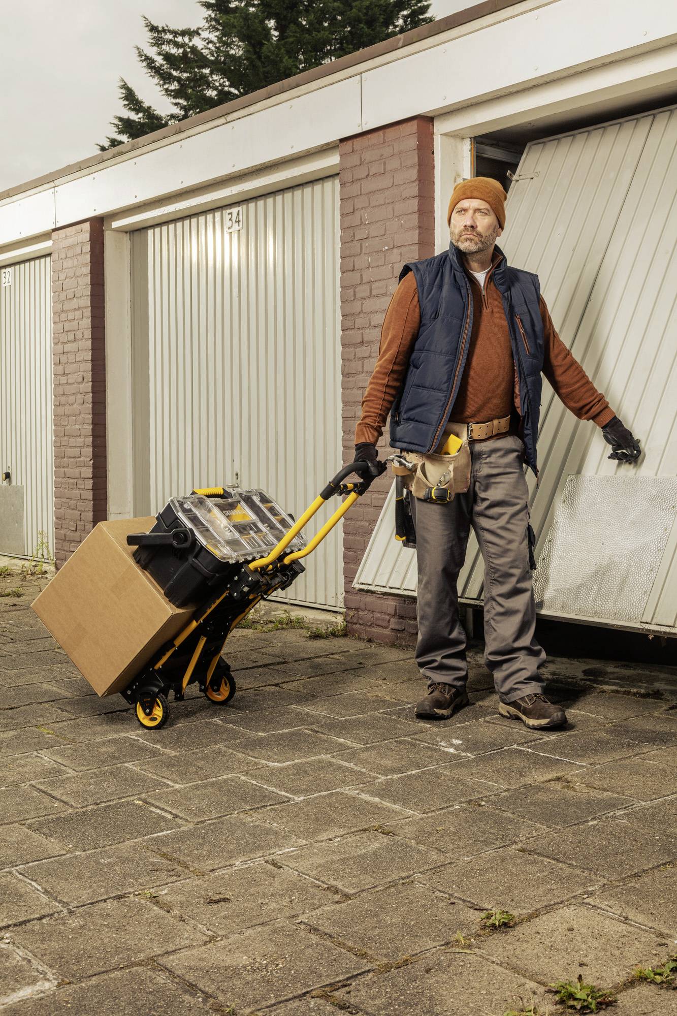 En man i arbetsklädsel står framför en öppen garageport och håller i en saxlyftvagn med en kartong och en verktygslåda.
