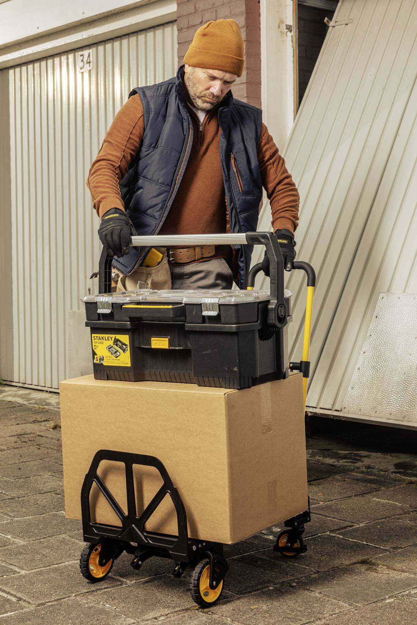En man transporterar en verktygslåda och en stor kartong på en handkärra framför en garagevägg.