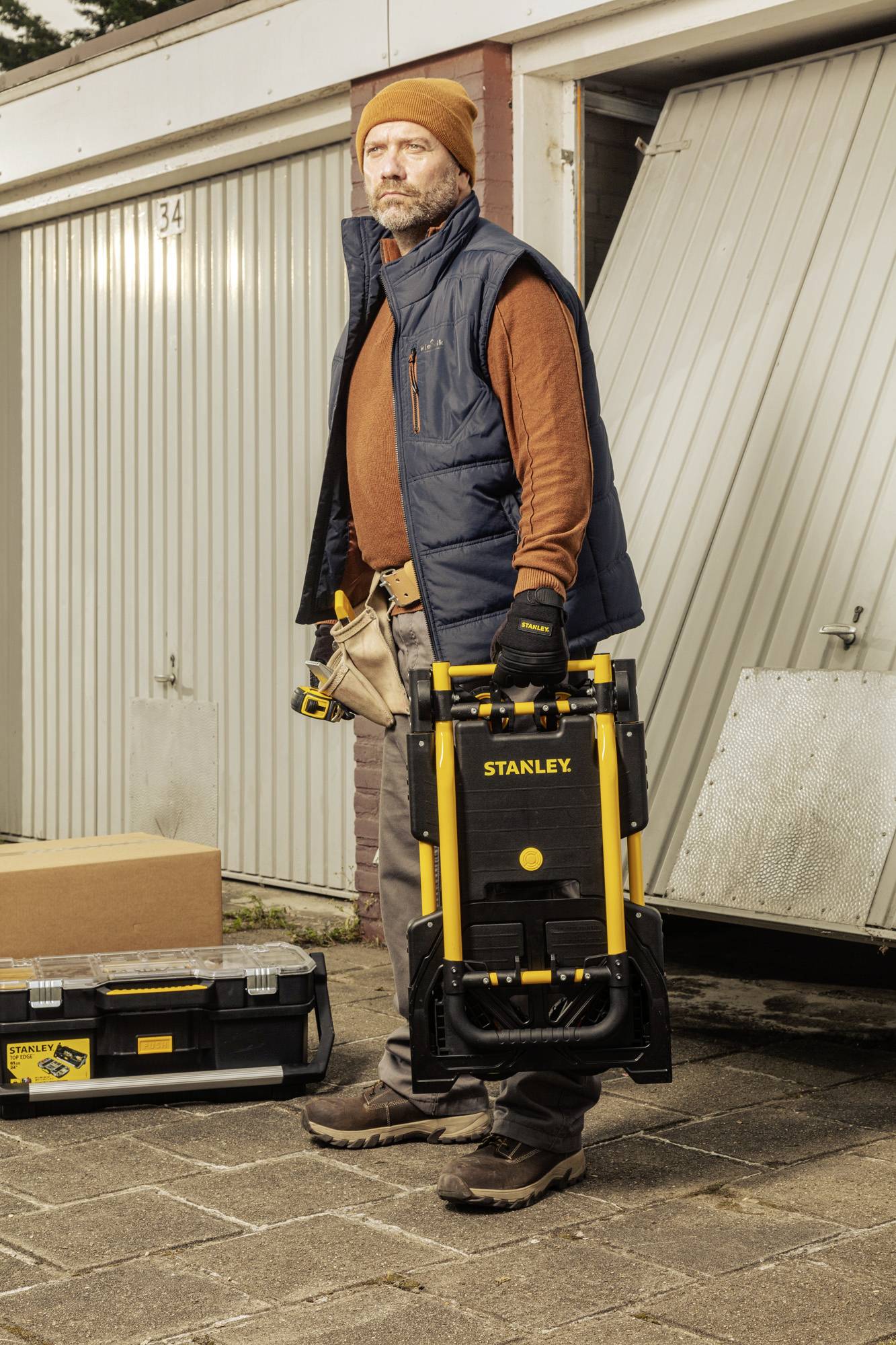 En man står framför en garage och håller i en ihopfälld vagn märkt 'Stanley'. Bredvid står en verktygslåda.
