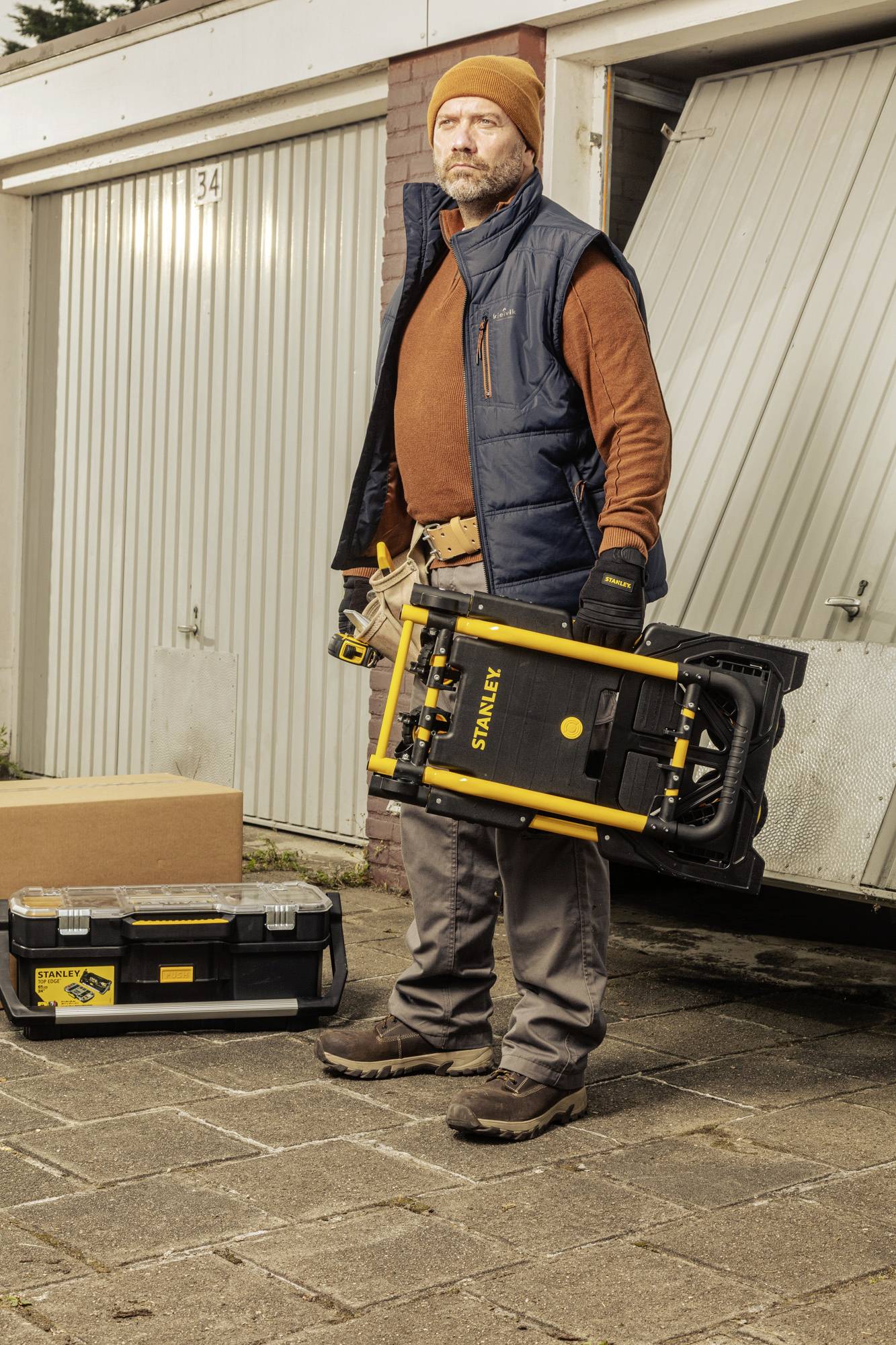 En man i arbetsklädsel står framför garagedörrar, håller i en gul verktygslåda och en svart låda.
