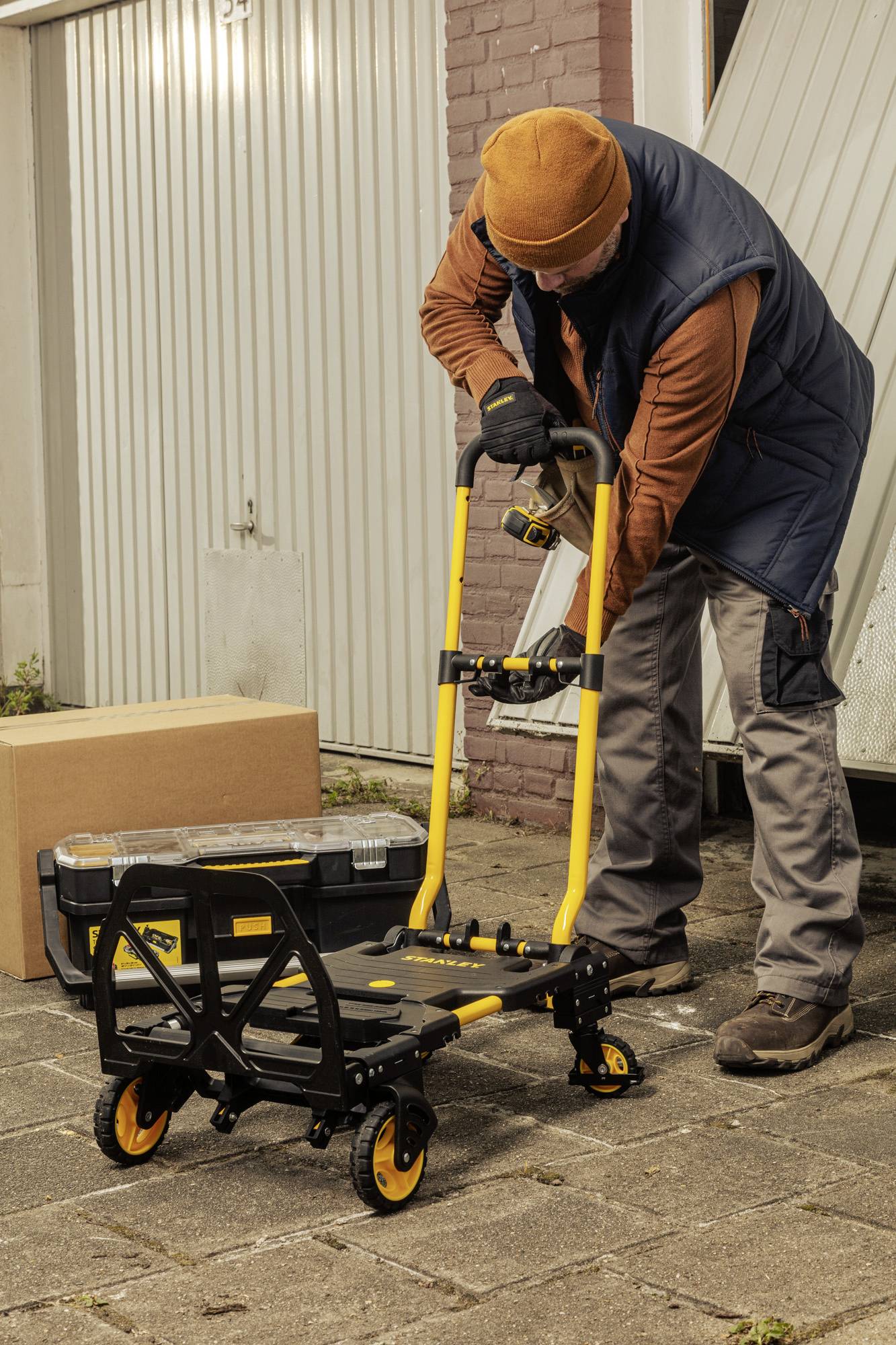 En person skjuter en gul handkärra, lastad med en verktygslåda, på en stenlagd gård framför ett garage.