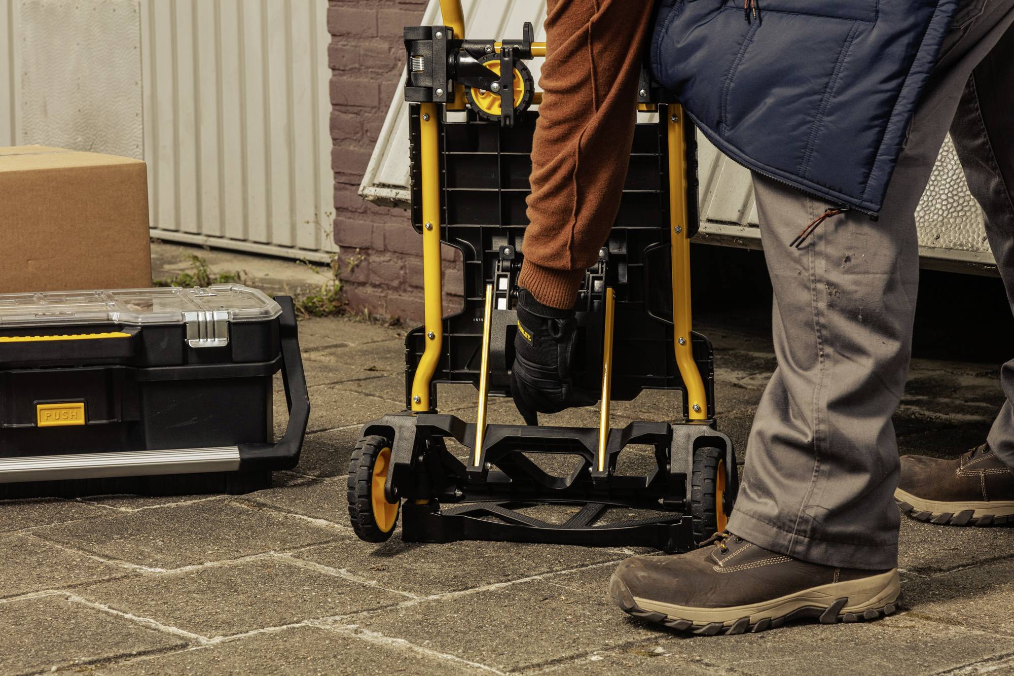En person ställer upp en hopfällbar vagn utomhus, omgiven av verktygslåda och paket.