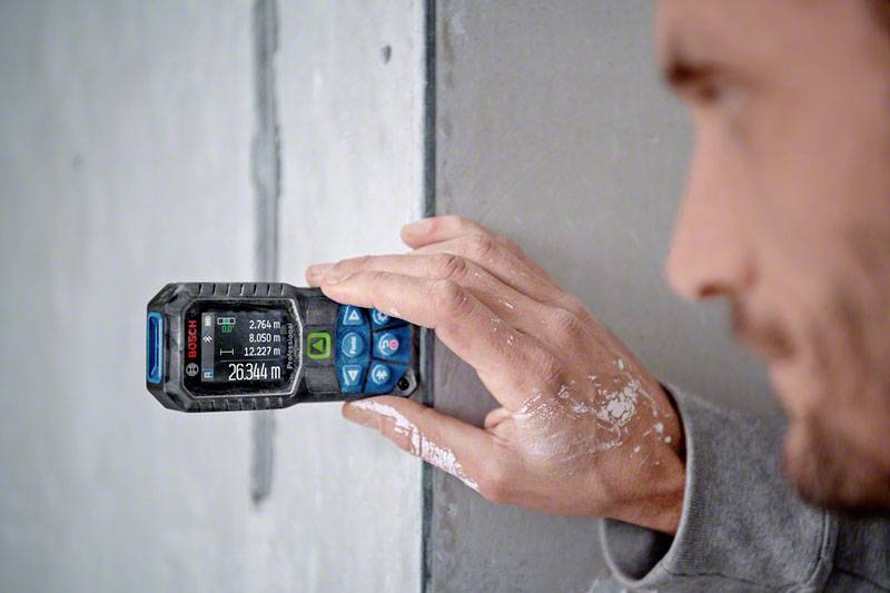 En person mäter längden på en betongvägg med en digital lasermätare. Enheten visar '2634 m'.