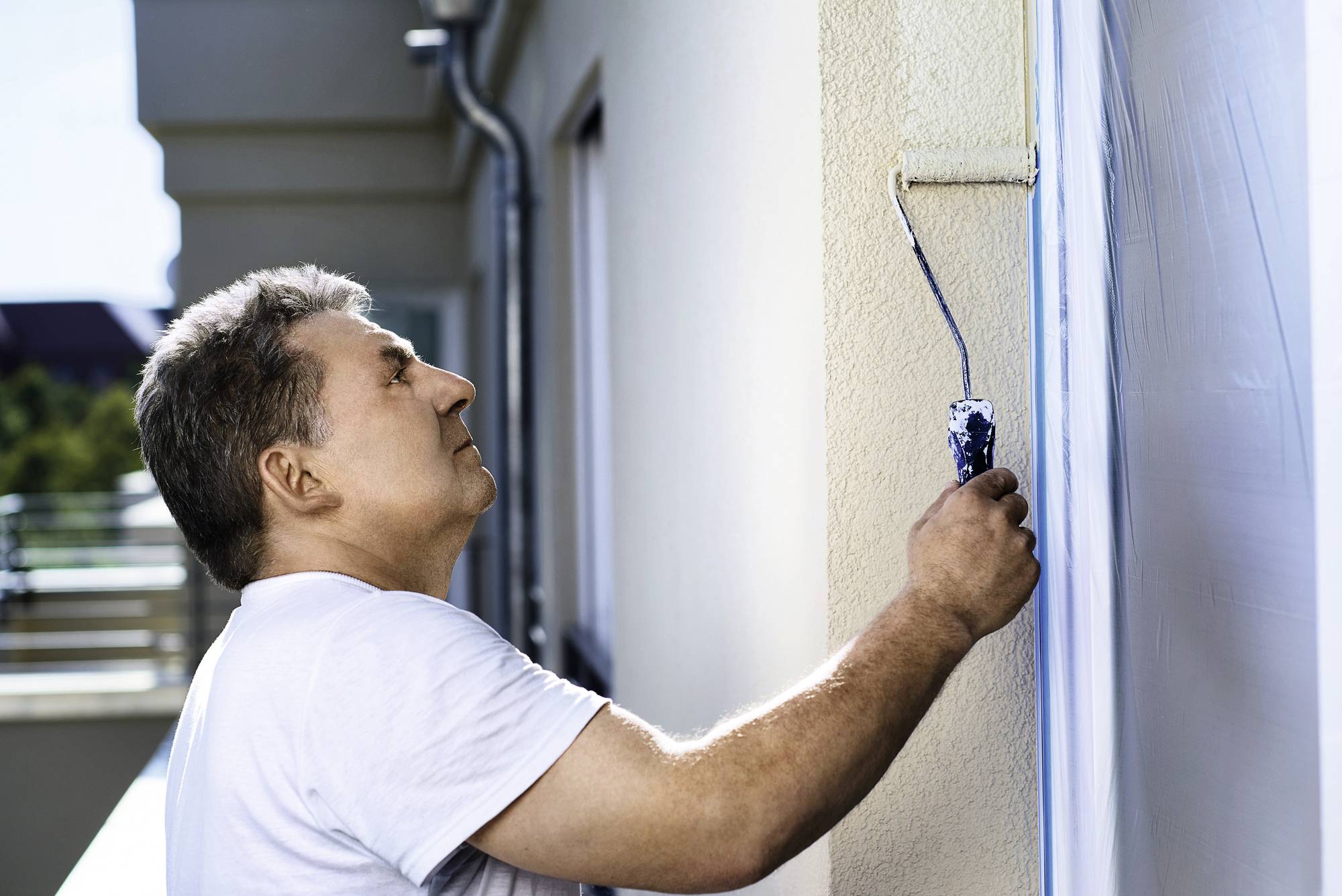 En man målar en husvägg med en vit färgrulle. Han är fokuserad på sitt arbete utomhus i dagsljus.