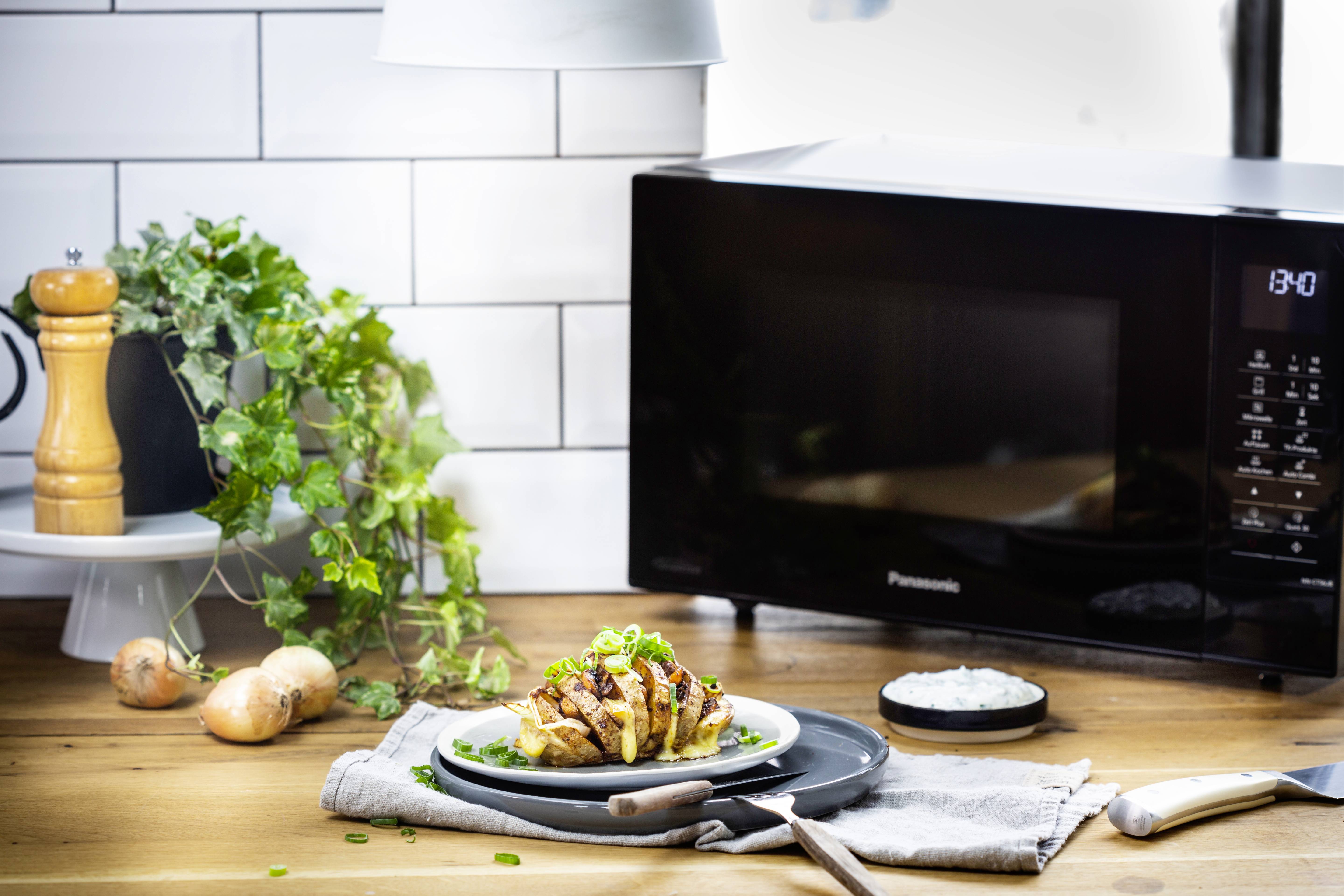 Uppställning på ett köksbord: Färska örter, hela lökar och en tallrik med potatis och skivat kött framför en mikrovågsugn.