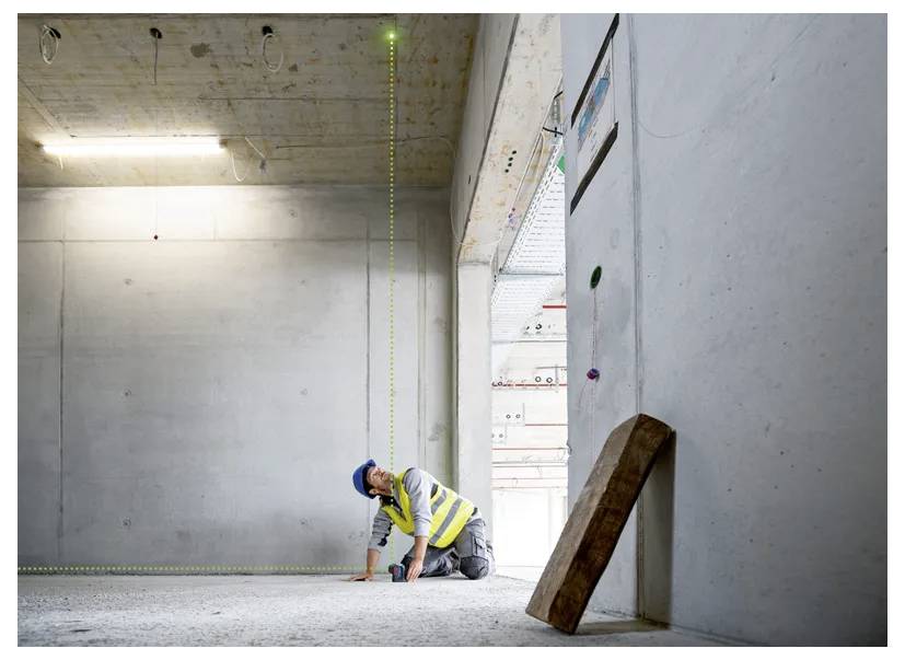 En byggnadsarbetare med hjälm och varselväst använder en lasernivå inuti en delvis färdigbyggd betongkonstruktion.