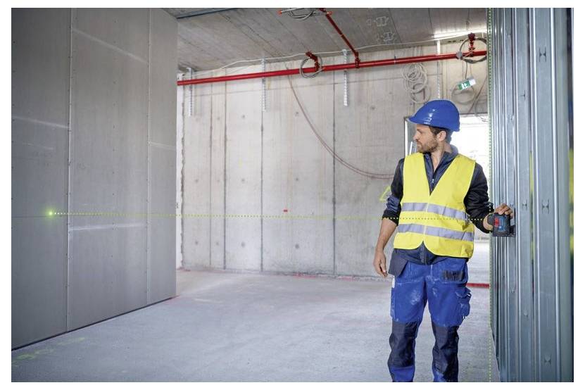En byggnadsarbetare i en säkerhetsjacka och hjälm använder ett lasernivåinstrument för att mäta i ett delvis färdigbyggt betongrum med synliga rör.