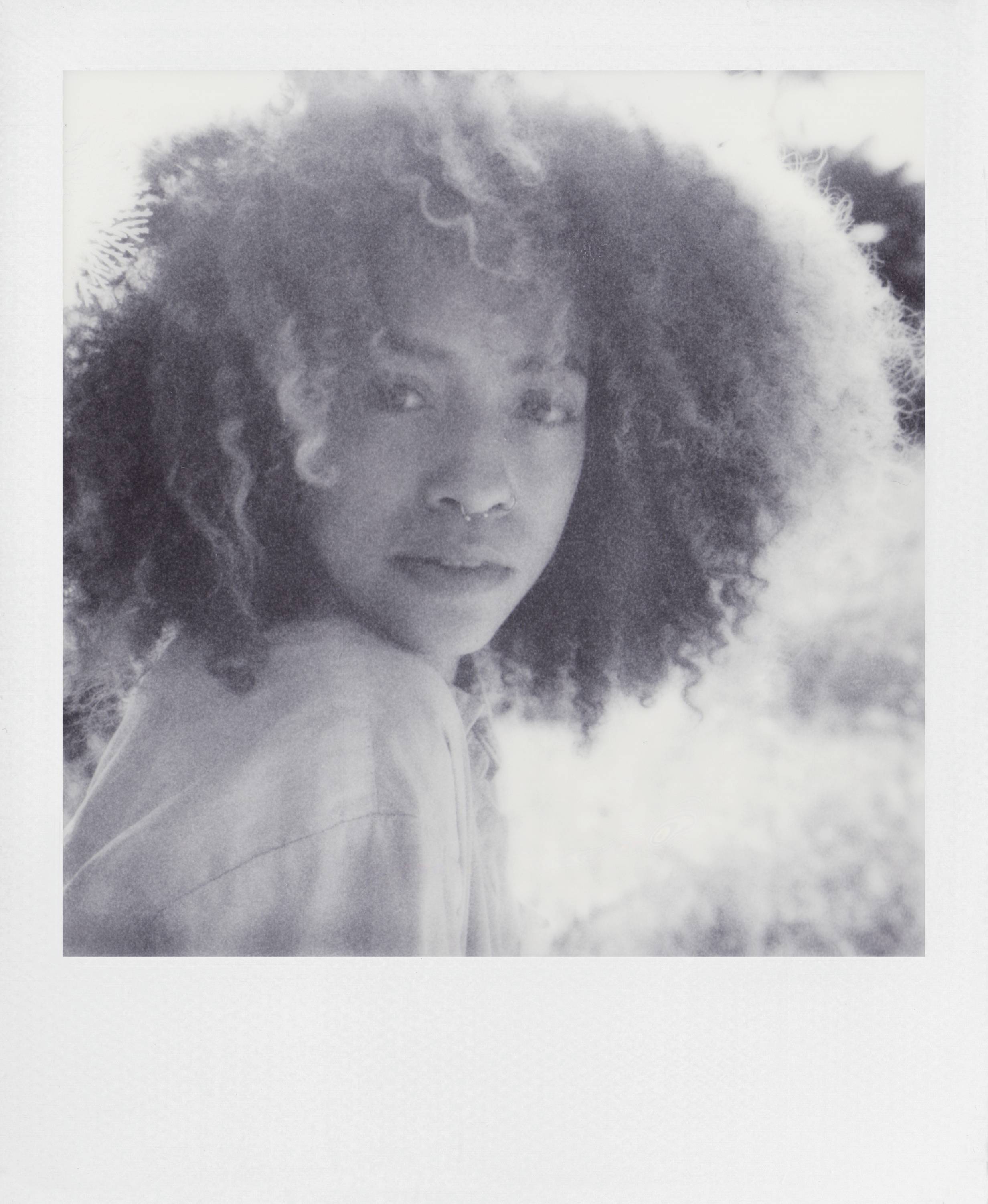 En person med lockigt hår blickar eftertänksamt in i kameran. Bakgrunden är oskarp och visar en naturlig miljö.