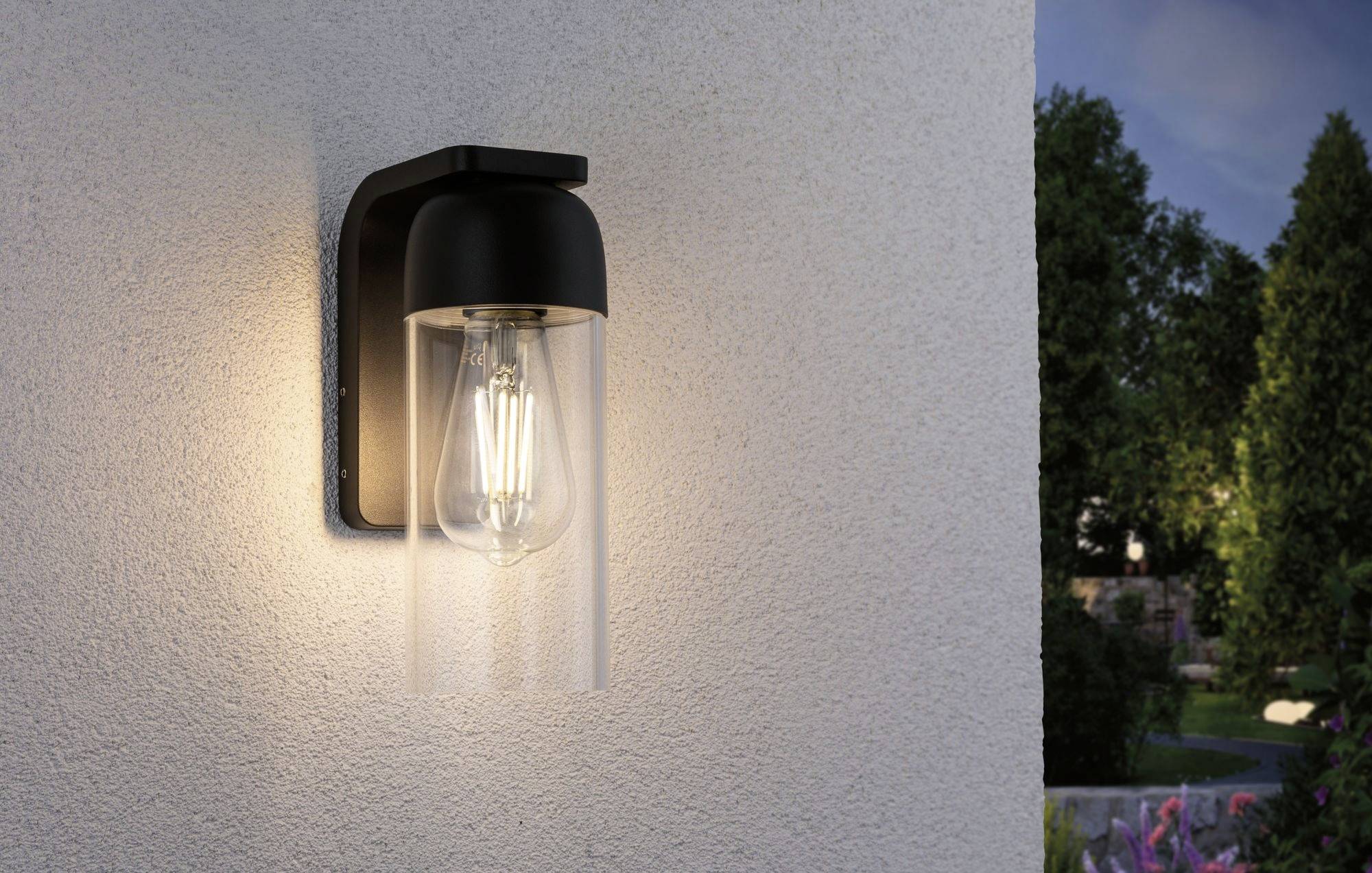 En modern utomhuslampa med svart hölje och glödlampa lyser mot en vägg, omgiven av en mörk trädgårdsbakgrund.