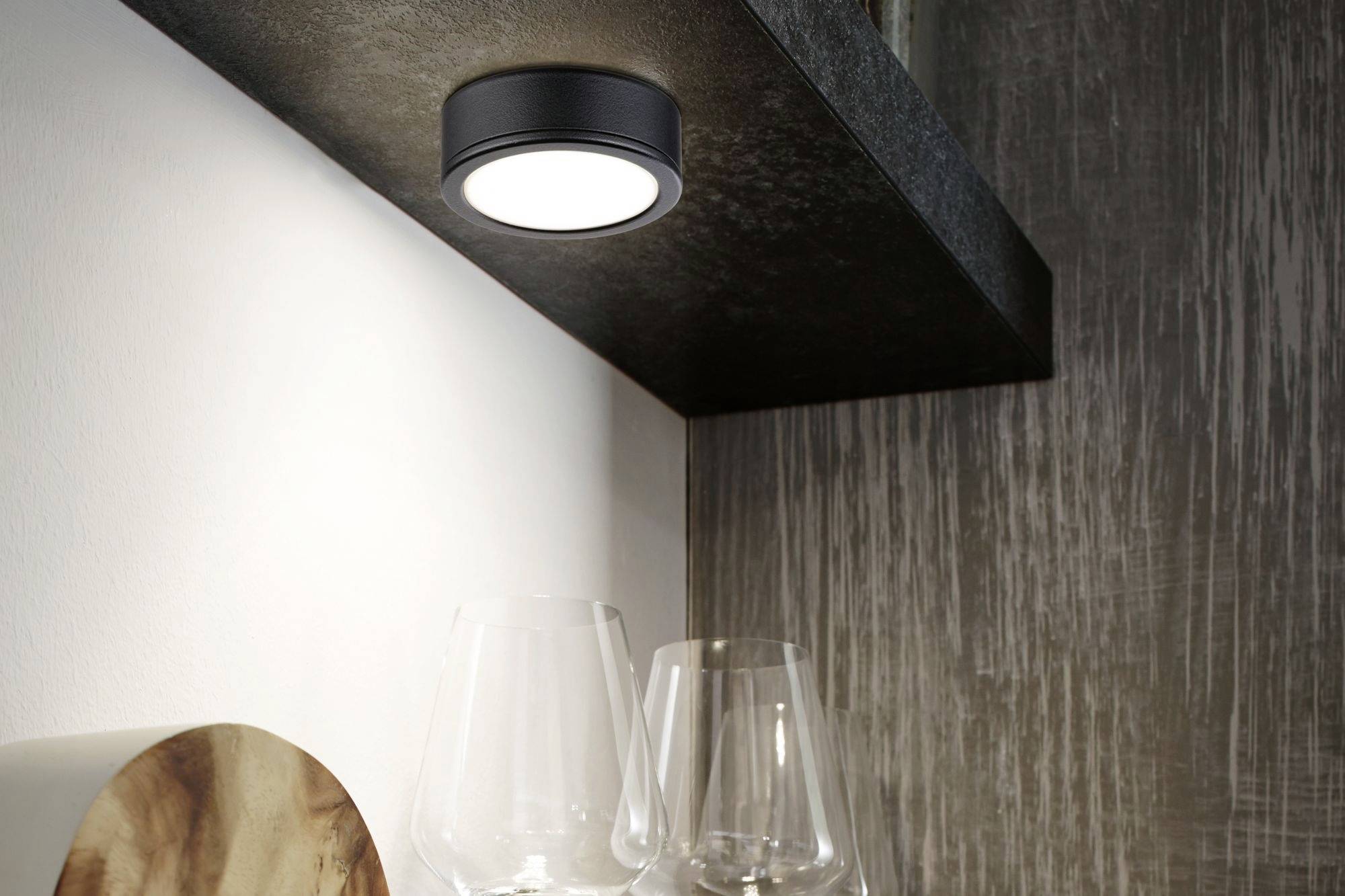 En rund lampa är monterad under en mörk hylla och lyser upp två tomma vinglas nedanför. Lyxig boendemiljö.