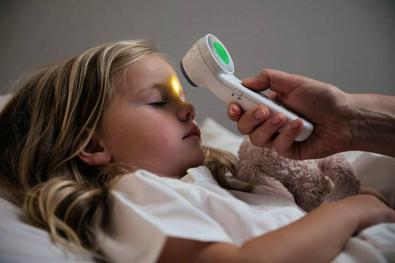 Ett barn ligger i sängen och sover, medan en hand för ett digitalt termometer mot dess panna för att mäta temperaturen.